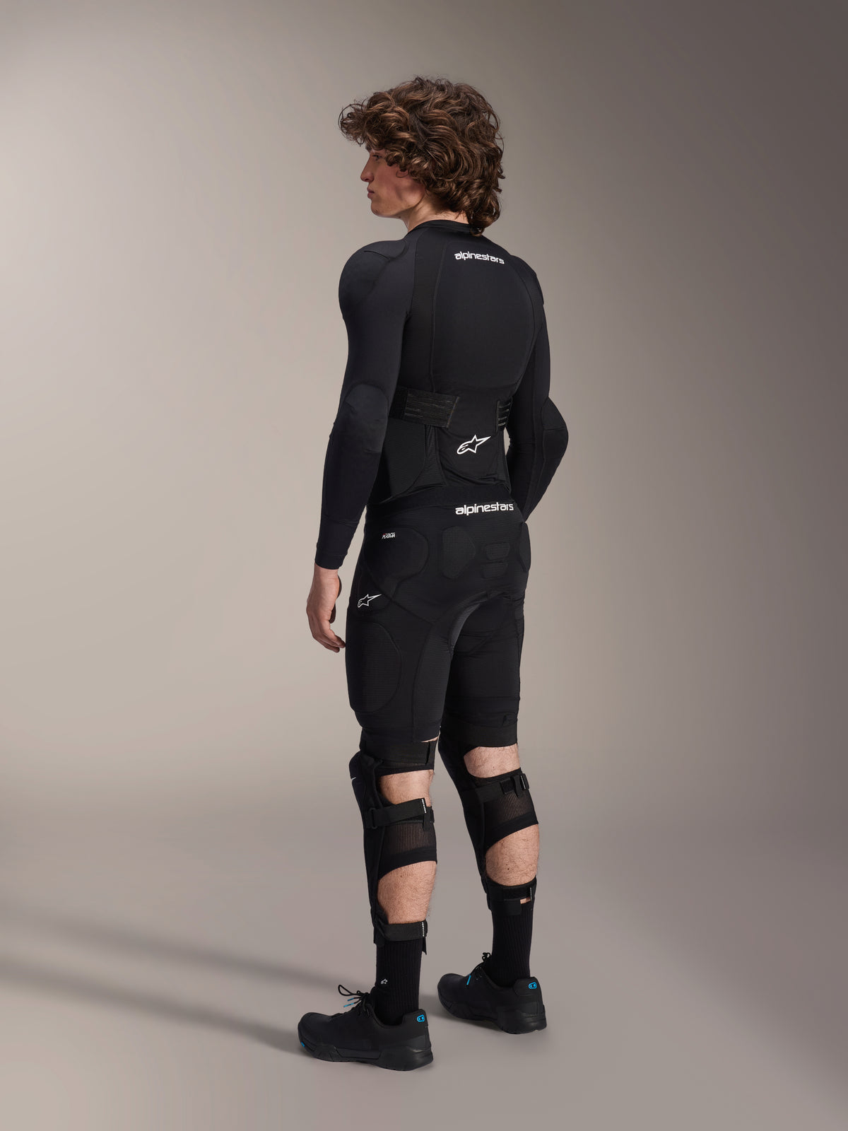 Model w ochraniaczach kolan i goleni Alpinestars A-Impact Plasma Pro, widok od tyłu, kolor czarno-biały, prezentujący potrójne zapięcie na paski i wentylowane panele z siatki, ubrany w pełny zestaw czarnej odzieży ochronnej MTB z koszulką z długim rękawem i spodenkami ochronnymi
