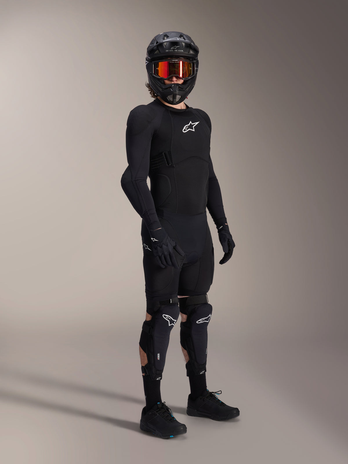 Alpinestars A-Impact Plasma Pro Knee Shin Protectors, czarno-białe, noszone przez rowerzystę w kompletnym stroju ochronnym MTB, zawierającym czarną zbroję z długim rękawem, czarne rękawice oraz kask typu full-face z pomarańczowymi goglami.