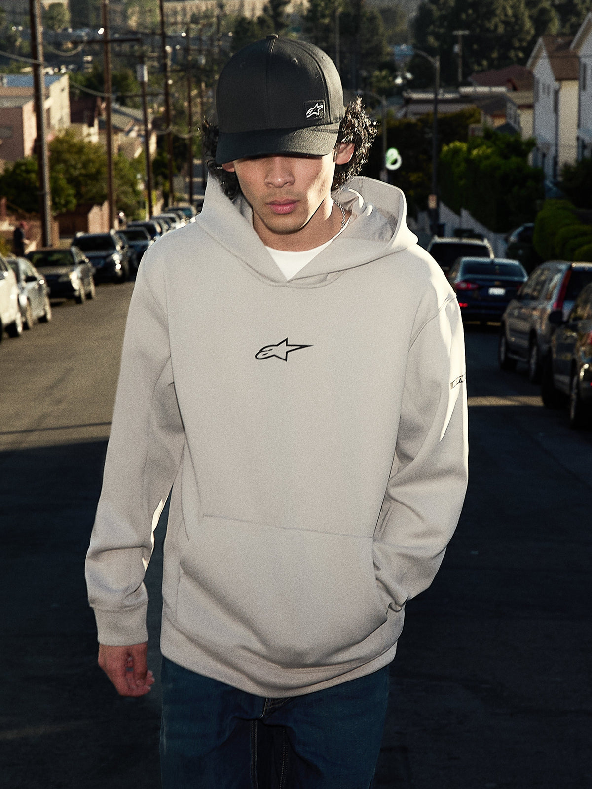 Bluza z kapturem Alpinestars Insight, codzienna odzież streetwearowa, kolor szary cementowy z czarnym logo Astars na piersi, fason typu pulower z kieszenią kangurką, prezentowana z czarną czapką na osiedlowej ulicy, nowoczesny design inspirowany sportami motorowymi