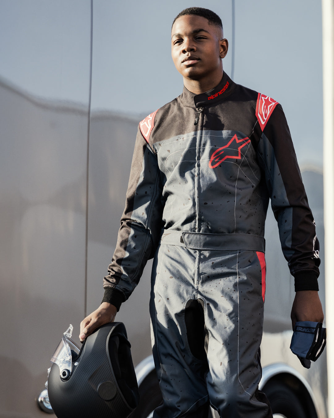 Kierowca gokartowy w kombinezonie Alpinestars KMX-9 V3 Graphic 6 w kolorze Anthracite Black Red, trzymający matowy czarny kask i rękawice, wyraźne czerwone logo Alpinestars, grafika z motywem rozprysków, plenerowa sceneria sportów motorowych.