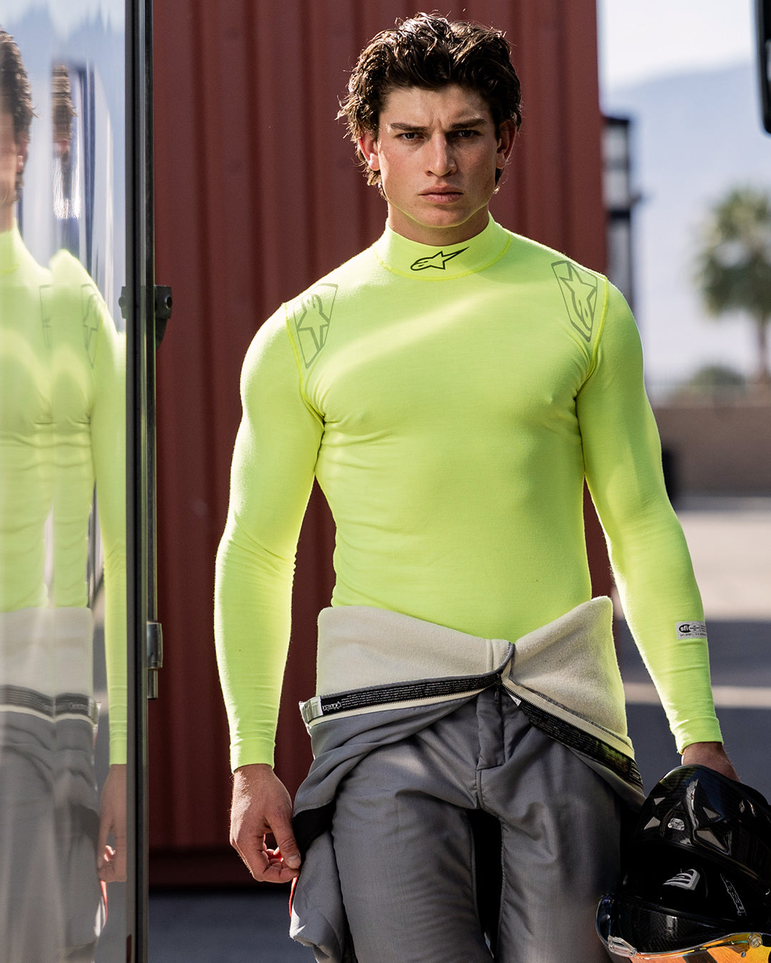 Alpinestars ZX Evo V3 Top - Long Sleeve, samochodowa bielizna termoaktywna, żółty neonowy, noszony przez kierowcę z szarym kombinezonem wyścigowym przewiązanym w pasie, trzymającego czarny kask, logo Alpinestars na kołnierzu i ramionach, techniczna odzież odprowadzająca wilgoć do sportów motorowych