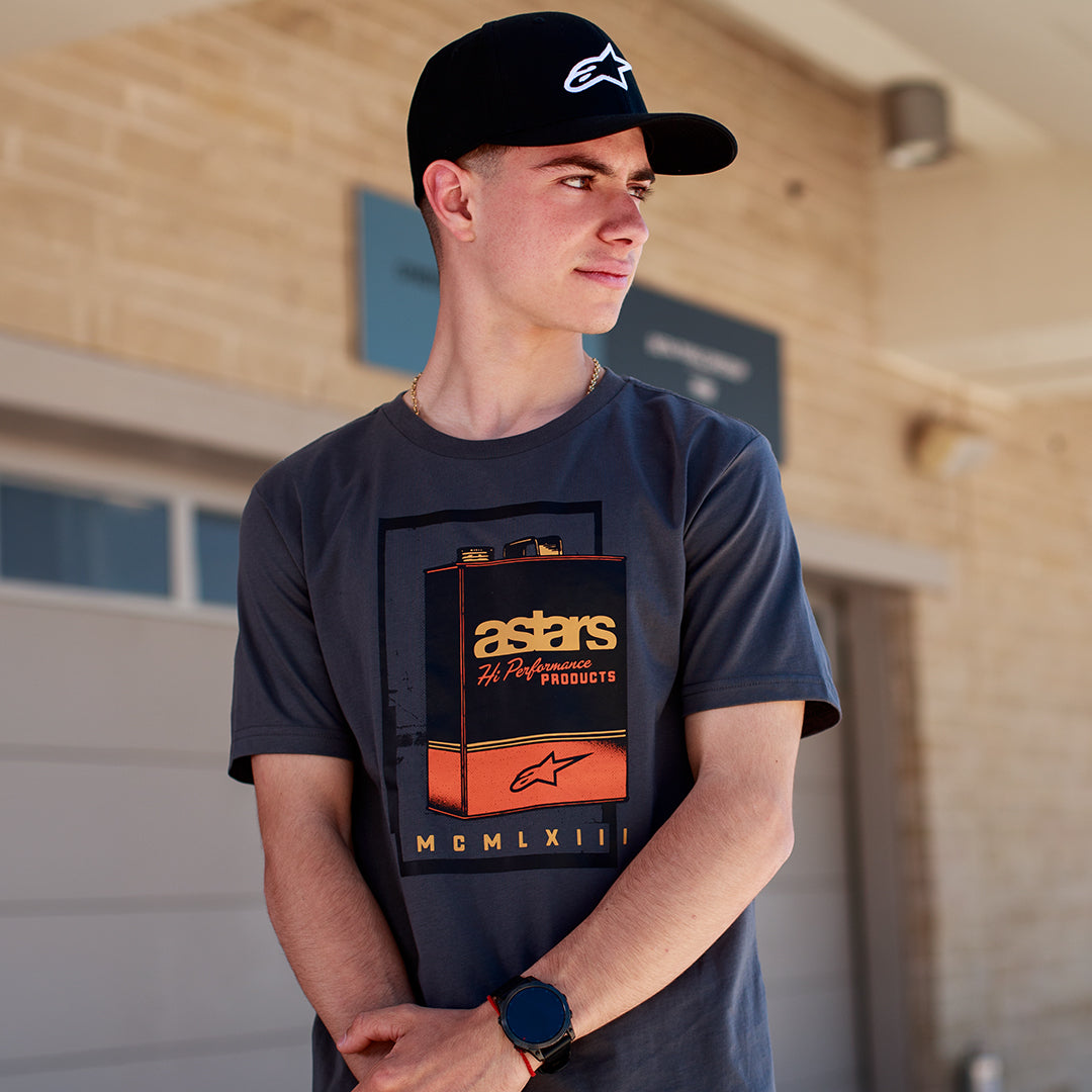Alpinestars Galun CSF Tee, koszulka z krótkim rękawem, szara, z przodu grafika z vintage kanistrem na benzynę, logo „astars” i rzymskie cyfry „MCMLXIII”, noszona przez mężczyznę w czarnej czapce z logo, codzienny styl motorsportowy