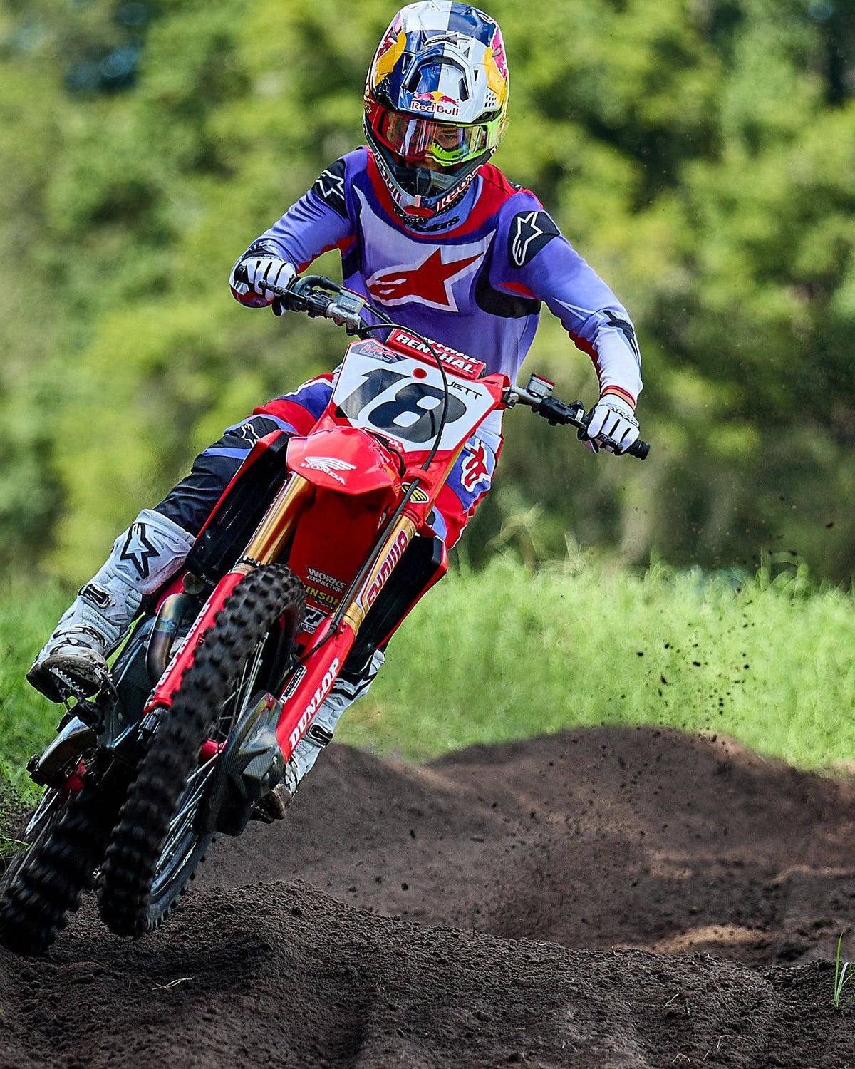 Motocyklista motocrossowy na czerwonym motocyklu Honda, ubrany w koszulkę Alpinestars 2025 Fluid Wurx w kolorze fioletowym i jasnoczerwonym, dopasowane spodnie, białe buty i kask Red Bull, pokonujący zakręt na torze ziemnym.