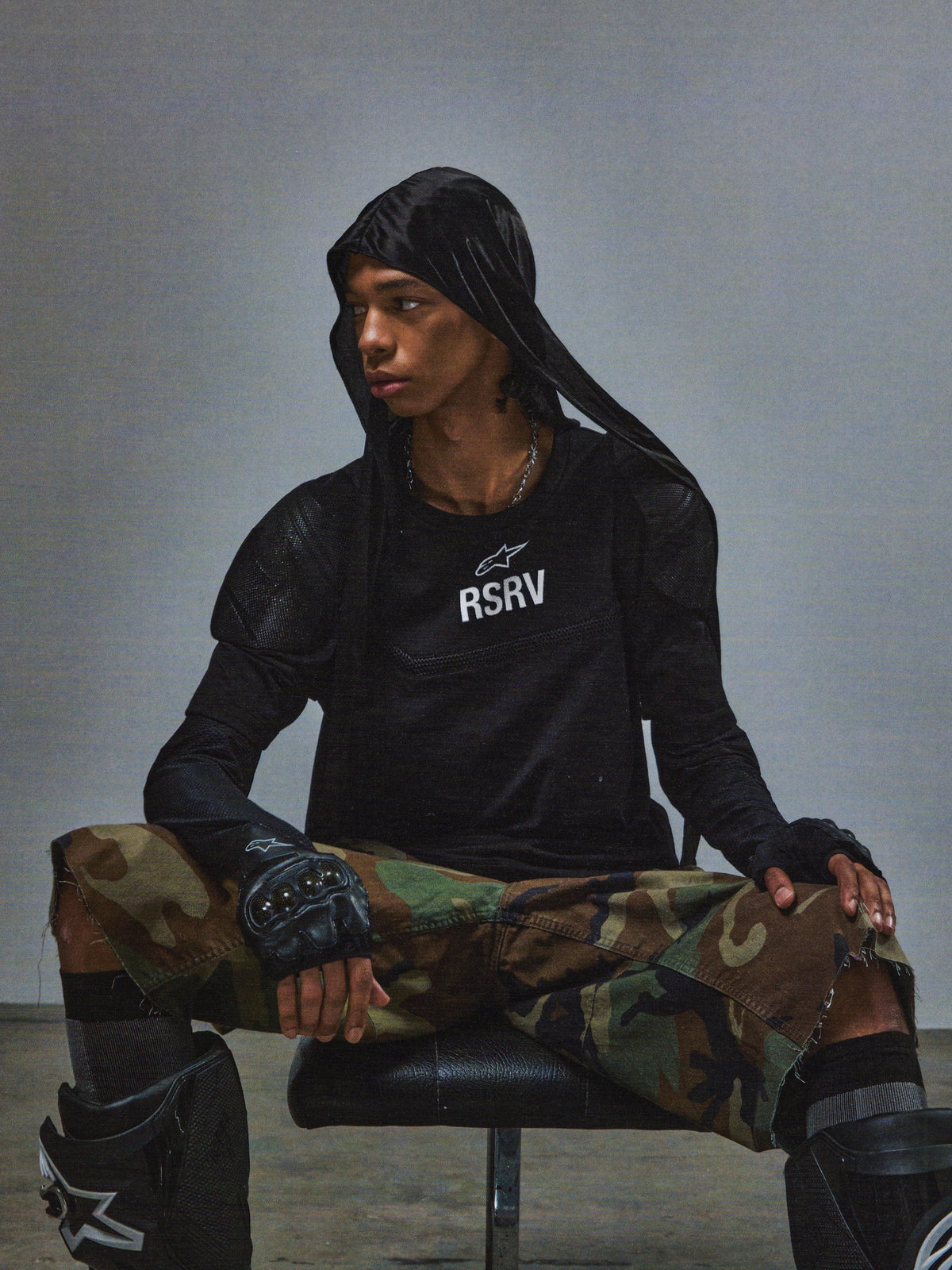 Alpinestars RSRV Nucleon SS Tee, czarna koszulka z nadrukiem RSRV, styl miejski i taktyczny, osoba w czarnej bluzie z kapturem, spodnie moro, rękawice bez palców, ochraniacze na kolana