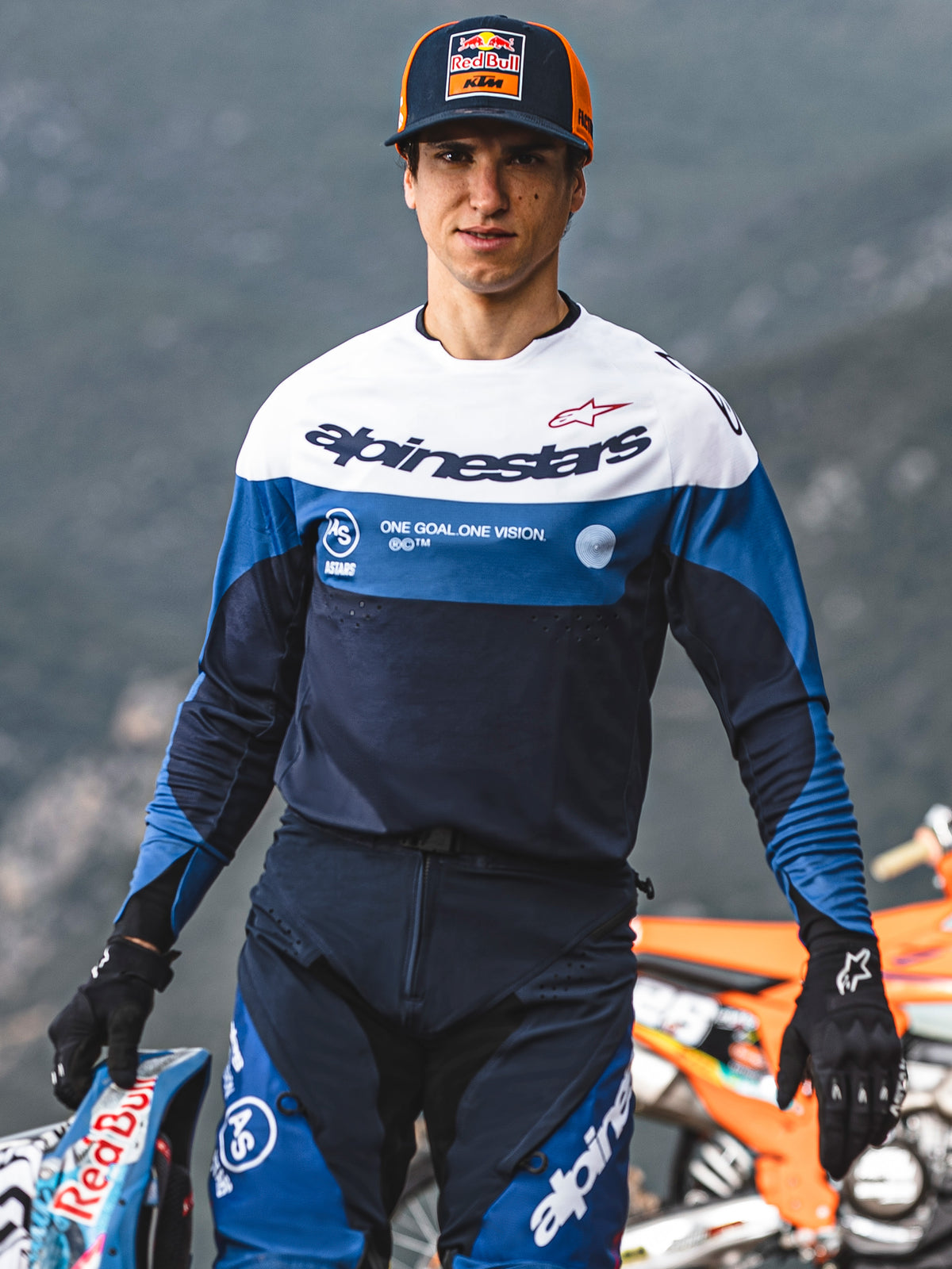Zawodnik motocrossowy w koszulce Alpinestars Pro-Dura w kolorach granatowym, jasnoniebieskim i białym, z logotypem Alpinestars i hasłem „One Goal. One Vision.”, w dopasowanym stroju sportowym i czapce Red Bull KTM, w plenerze.