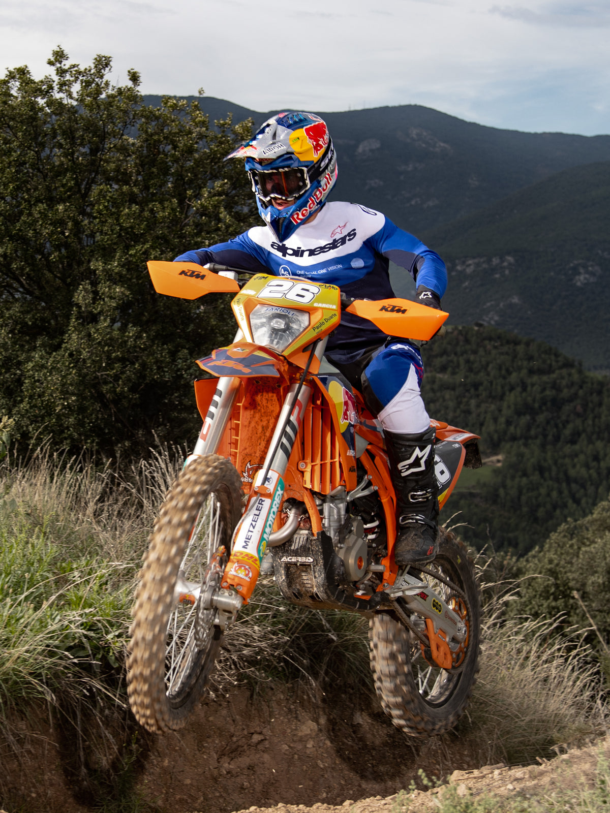 Motocyklista enduro na pomarańczowym motocyklu terenowym, koszulka Alpinestars Pro-Dura w kolorze granatowym, jasnoniebieskim i białym, czarno-białe spodnie oraz czarne buty Alpinestars, pokonujący trudny szlak z górami i drzewami w tle.