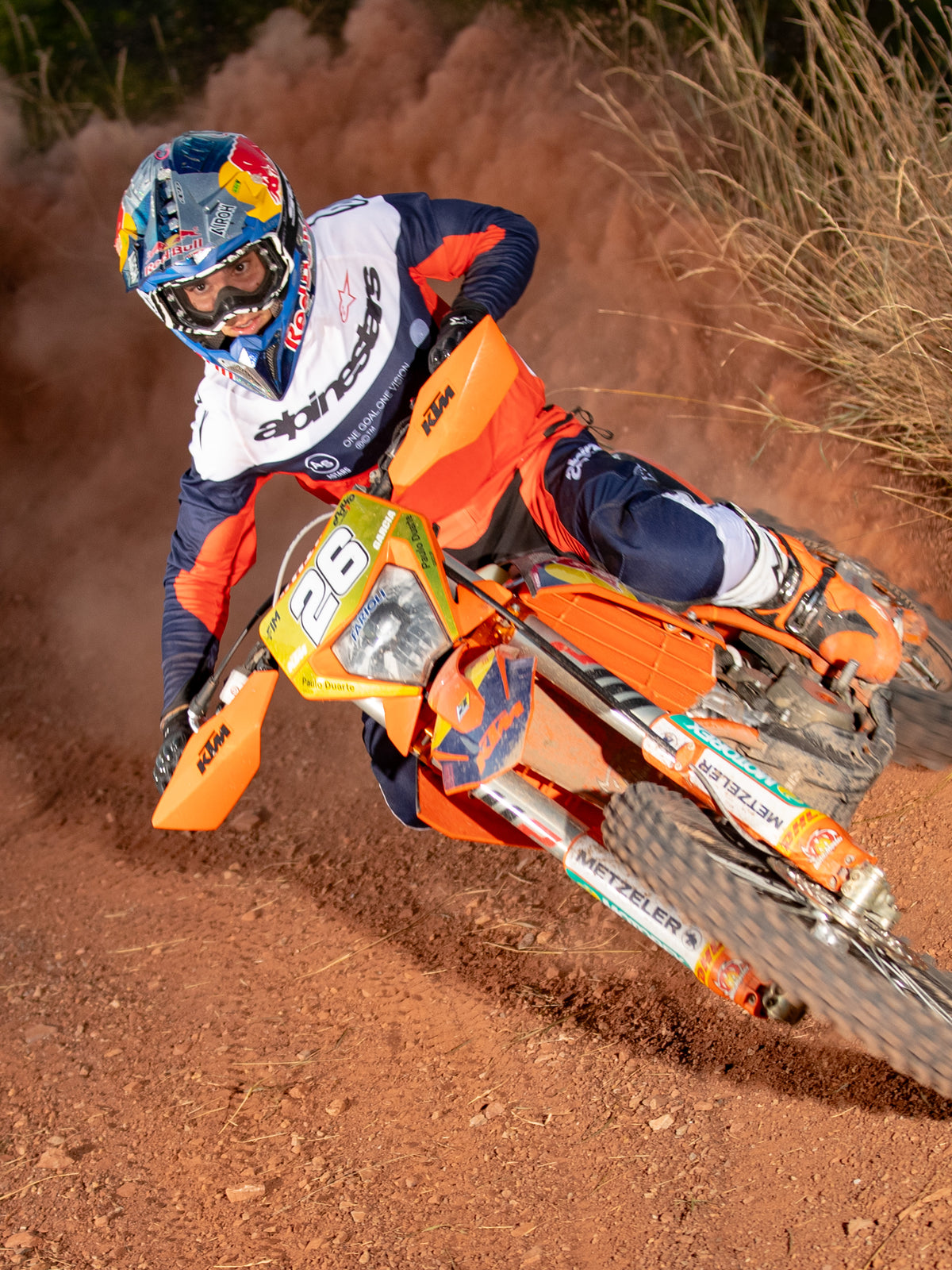 Motocyklista enduro w akcji na pomarańczowym motocyklu terenowym, ubrany w koszulkę Alpinestars Pro-Dura w kolorach granatowym, jaskrawopomarańczowym i białym, charakteryzującą się lekką i oddychającą konstrukcją z wyraźnym brandingiem dla wydajności w terenie.