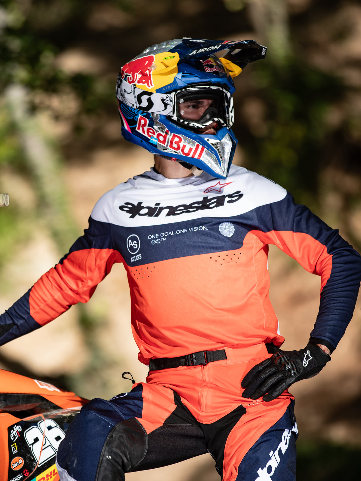 Motocyklista motocrossowy w koszulce Alpinestars Pro-Dura w kolorach granatowym, intensywnym pomarańczowym i białym, z wyraźnym brandingiem na piersi i laserowymi perforacjami, w kasku wyścigowym Red Bull, profesjonalny sprzęt do enduro i off-road.