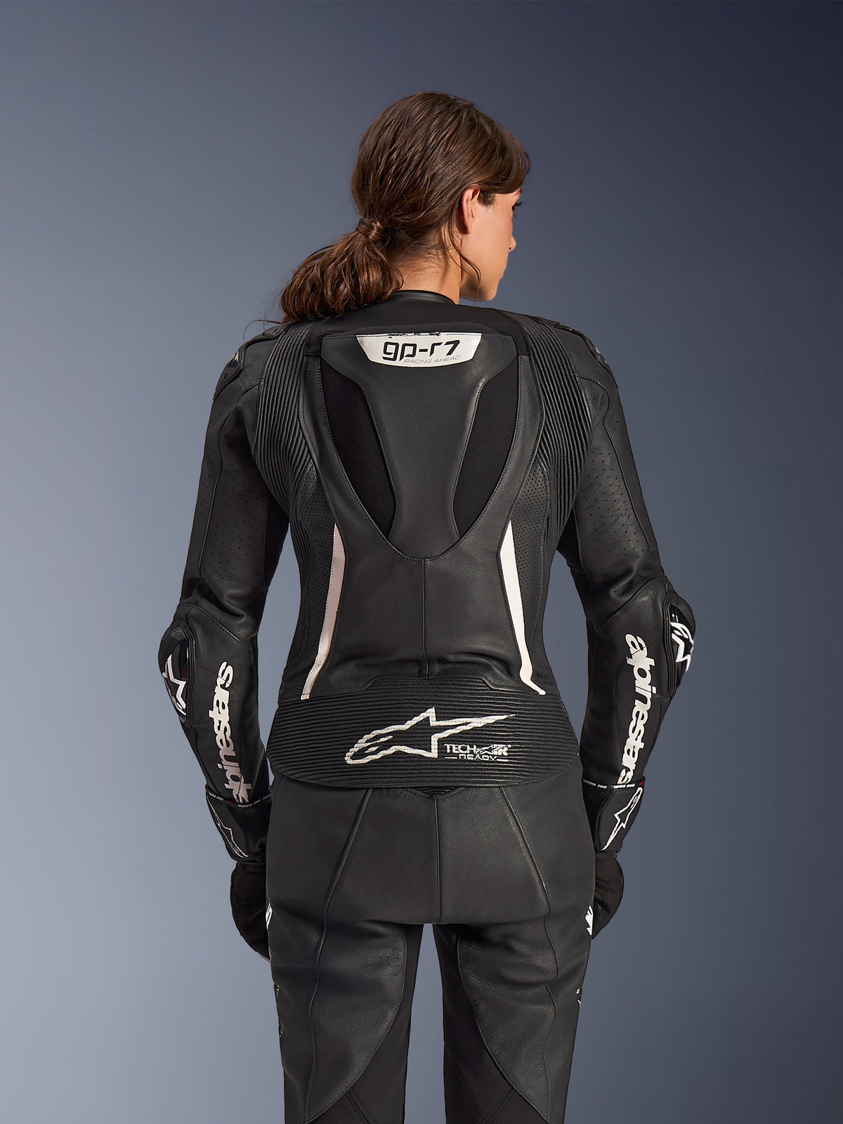 Dwuczęściowy skórzany kombinezon Alpinestars Stella GP-R7, damski kombinezon motocyklowy, czarny z białymi akcentami, widok z tyłu przedstawiający garb aerodynamiczny Supertech R10 z logo GP-R7, oznaczenie Tech-Air Ready w dolnej części pleców, białe napisy Alpinestars na rękawach oraz elastyczne panele harmonijkowe
