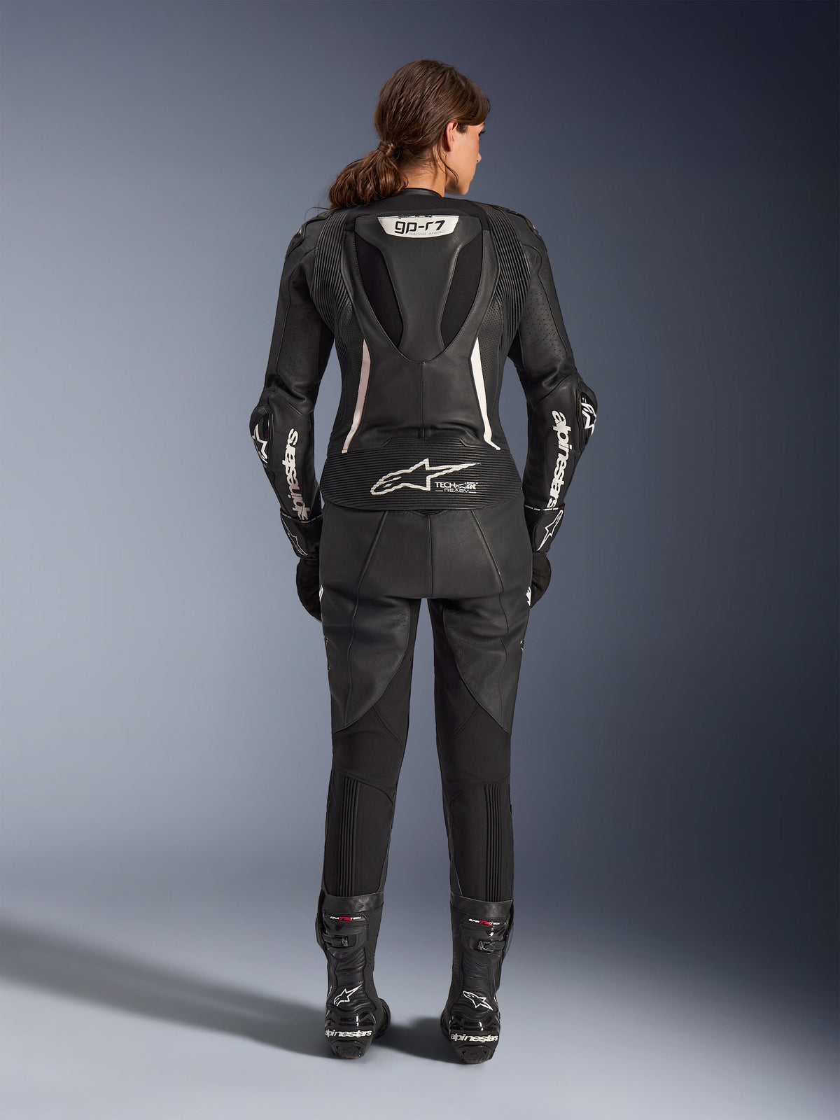 Alpinestars Stella GP-R7 2-częściowy skórzany kombinezon, damski kombinezon motocyklowy, czarny z białymi akcentami, widok z tyłu pokazujący garb aerodynamiczny GP-R7 i oznaczenie Tech-Air Ready, białe logo Alpinestars na rękawach, elastyczne panele harmonijkowe, zestawiony z czarnymi butami wyścigowymi