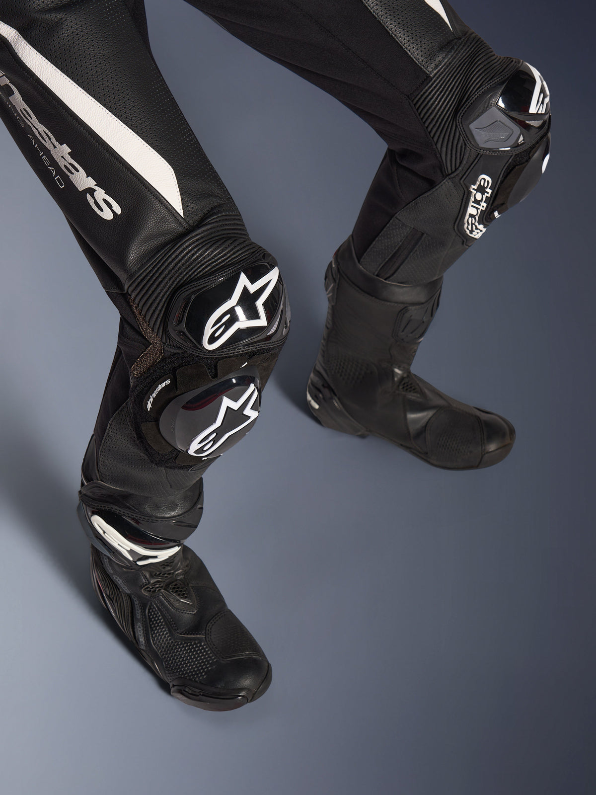 Detal nóg 2-częściowego skórzanego kombinezonu Alpinestars Stella GP-R7, czarny z białymi akcentami, ochraniacze kolan GP DFS RR i ślizgacze z logo Alpinestars, czarne buty wyścigowe, zaprojektowany do jazdy na torze