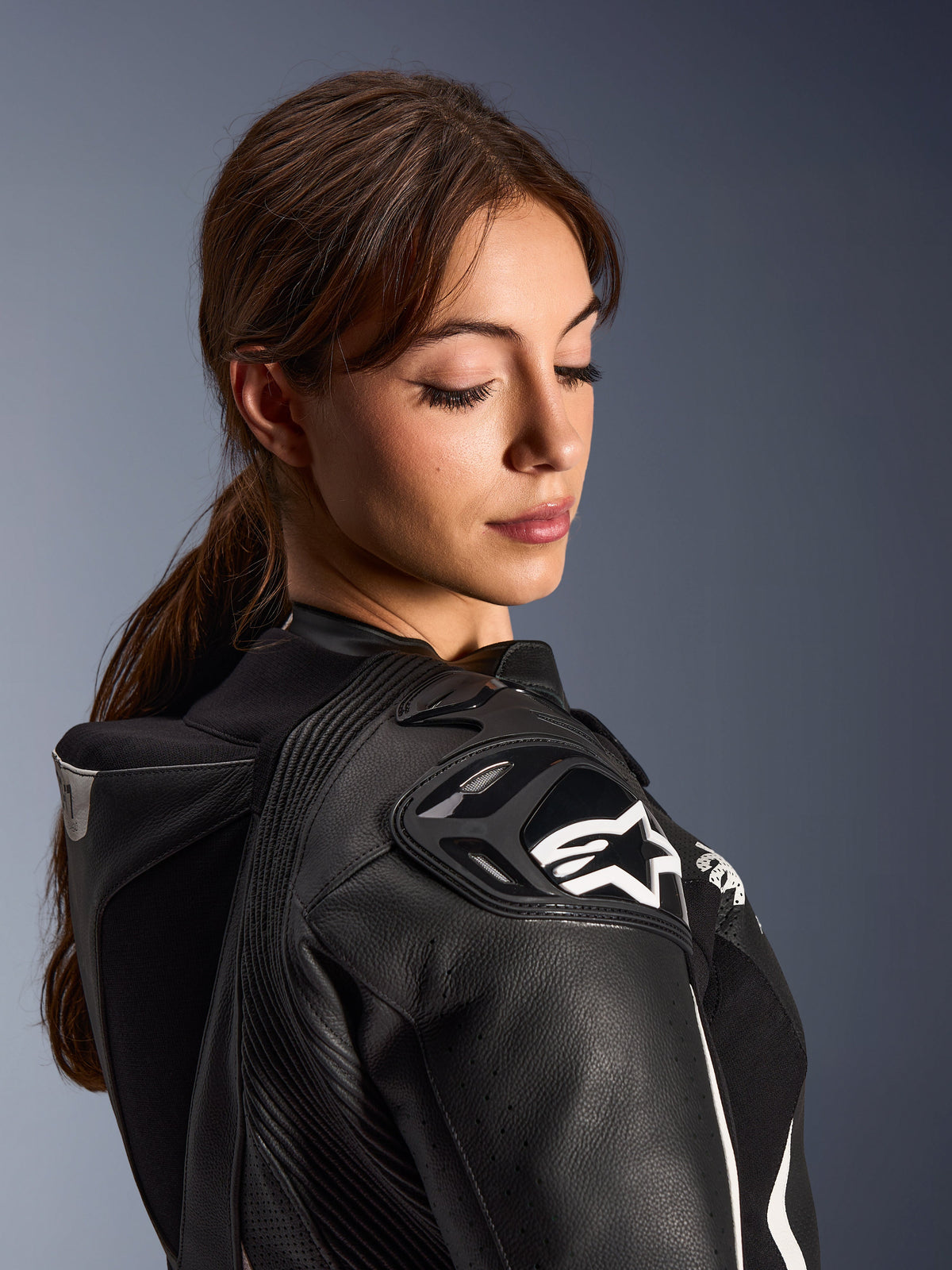 Kobieta w dwuczęściowym skórzanym kombinezonie Alpinestars Stella GP-R7, zbliżenie na ramię z czarnej skóry z ochraniaczem DFS RR i białym logo gwiazdy, zintegrowany garb aerodynamiczny i perforowane detale