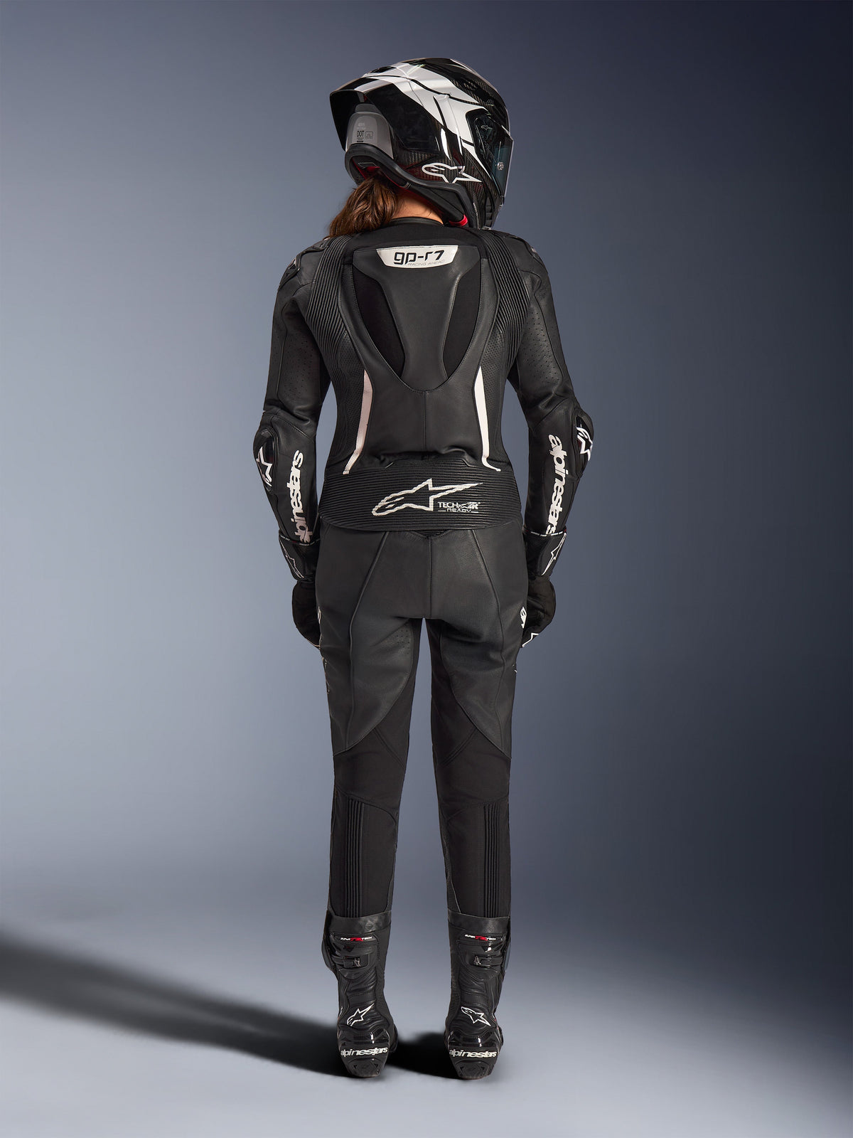 Damski dwuczęściowy skórzany kombinezon Alpinestars Stella GP-R7, czarny z białymi akcentami, widok z tyłu prezentujący garb aerodynamiczny Supertech R10 i logo Tech-Air Ready, noszony z pasującym kaskiem integralnym, rękawicami wyścigowymi i skórzanymi butami do jazdy na torze i sportowej