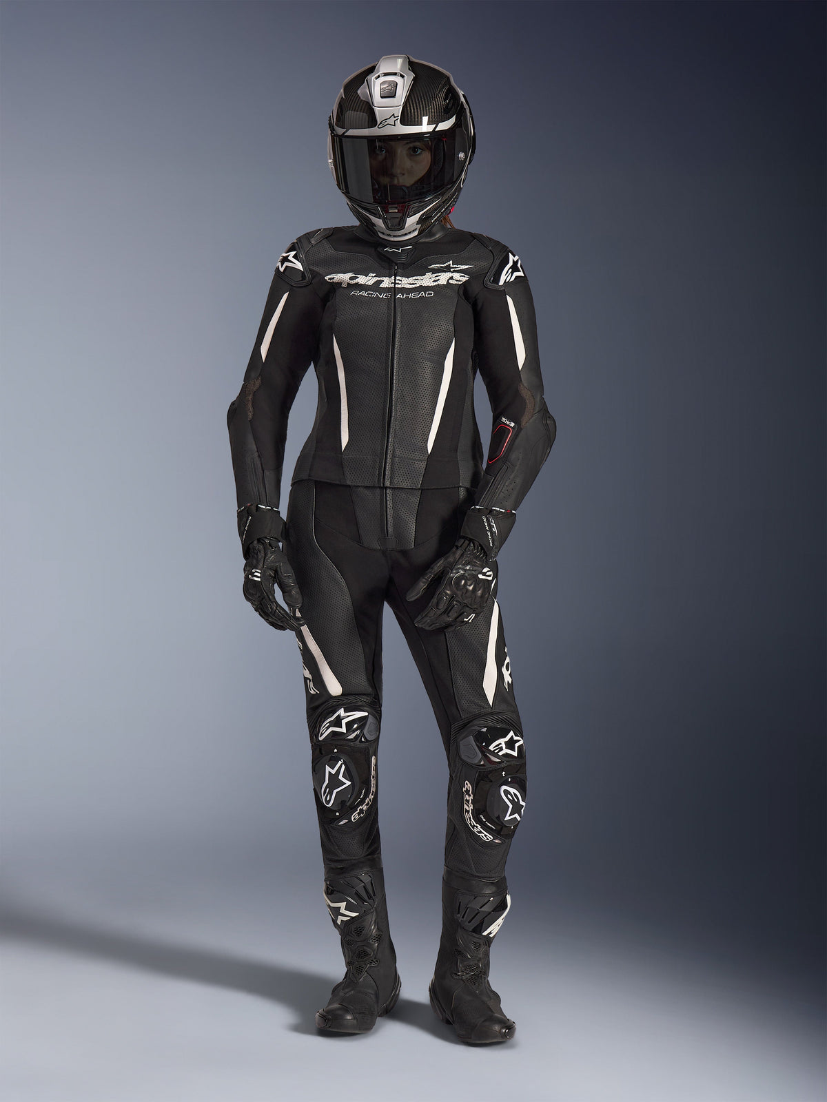 Motocyklistka w dwuczęściowym skórzanym kombinezonie Alpinestars Stella GP-R7, kolor czarny z białymi akcentami, kask integralny z włókna węglowego, czarne rękawice wyścigowe i buty, perforowane panele skórzane i zintegrowane ochraniacze, profesjonalny strój motocyklowy na tor i ulicę