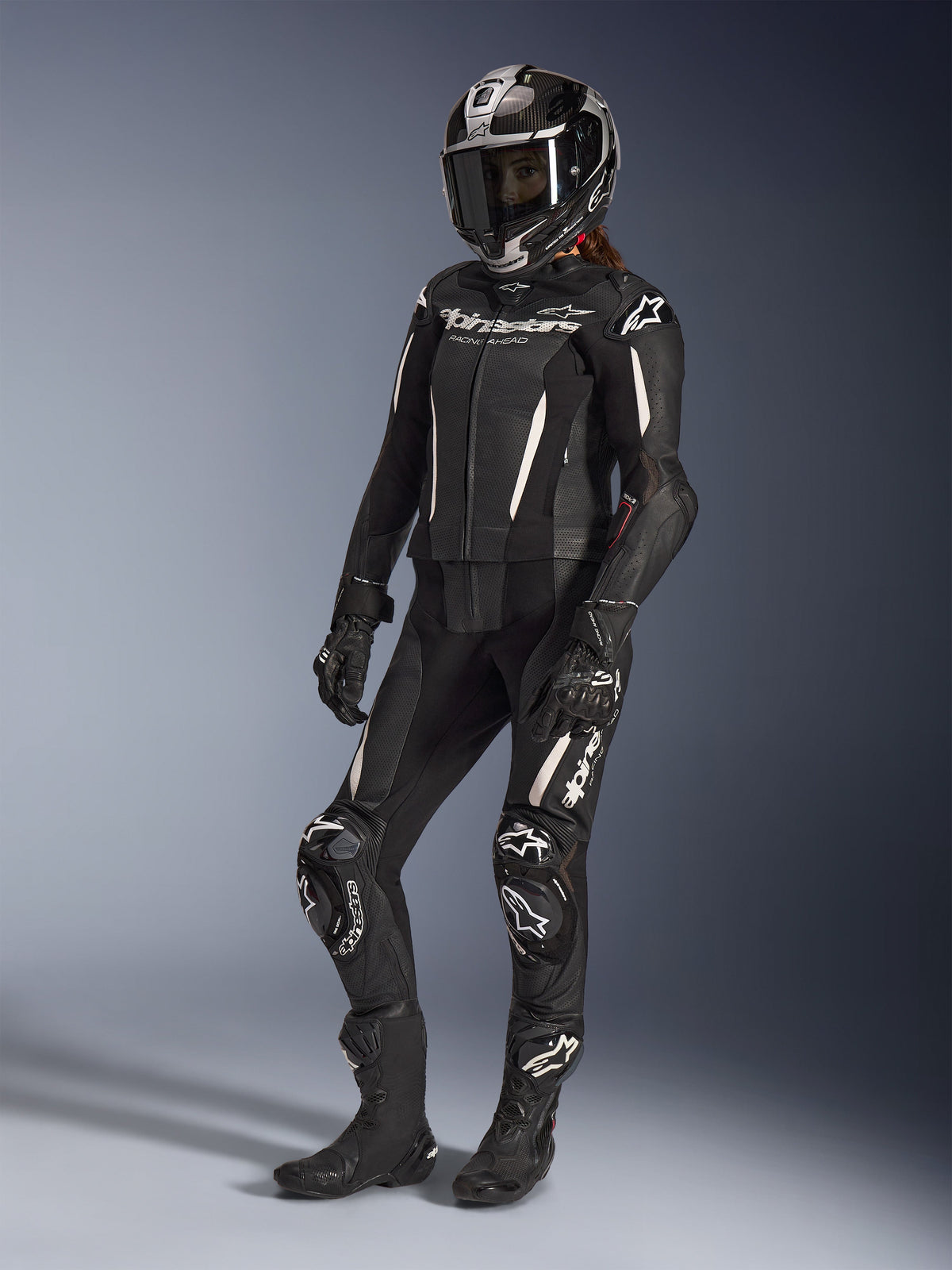 Alpinestars Stella GP-R7 2-Piece Leather Suit, damski kombinezon motocyklowy, czarny z białymi akcentami, noszony przez motocyklistkę w pełnym kasku wyścigowym, czarnych rękawicach i butach, perforowane panele skórzane i białe logo Alpinestars, profesjonalna odzież wyścigowa i sportowa