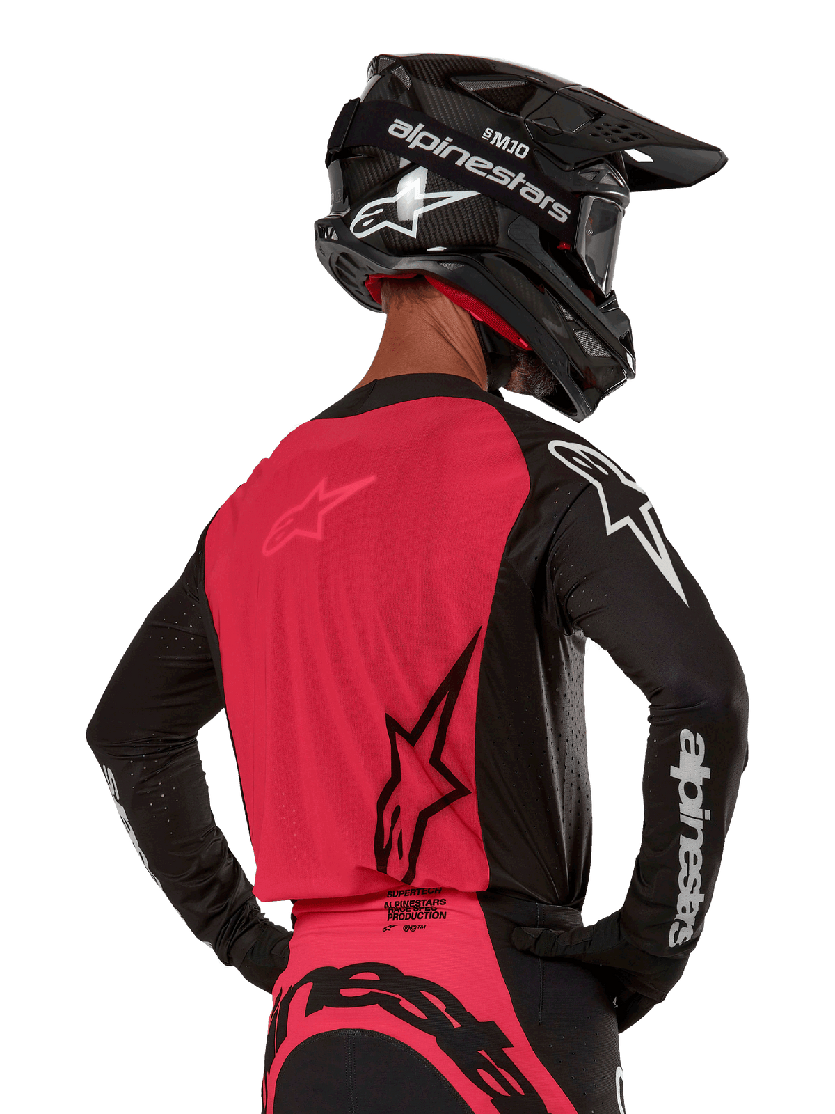 Alpinestars 2024 Supertech Ward Jersey, koszulka motocrossowa, czarno-czerwona (Red Berry), widok z tyłu prezentujący czerwony panel z siatki i czarne rękawy z białym brandingiem, zestawiona z czarnym kaskiem z włókna węglowego i rękawicami, sportowy krój wyścigowy do jazdy terenowej