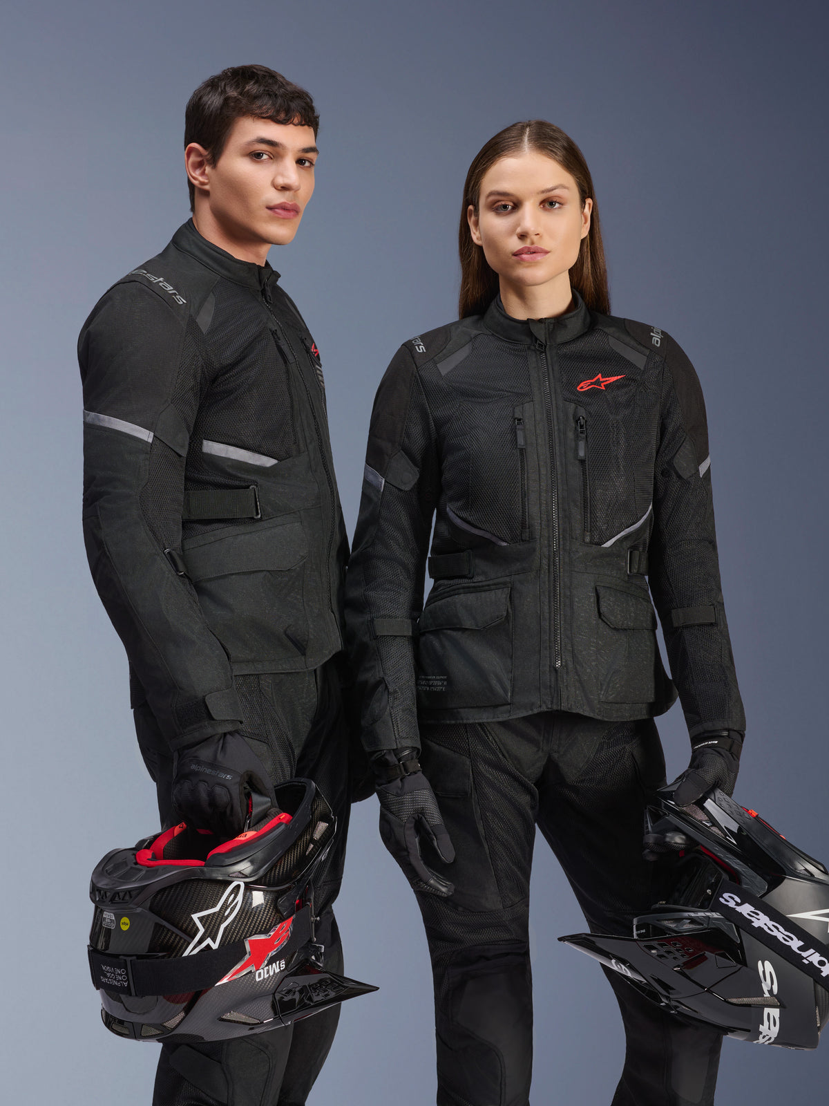 Model i modelka ubrani w czarne kurtki Alpinestars Stella Andes Air Drystar®, turystyczna odzież motocyklowa adventure z czerwonym logo gwiazdy, siatkowe panele wentylacyjne i techniczna konstrukcja tekstylna, trzymający kaski adventure, zdjęcie studyjne