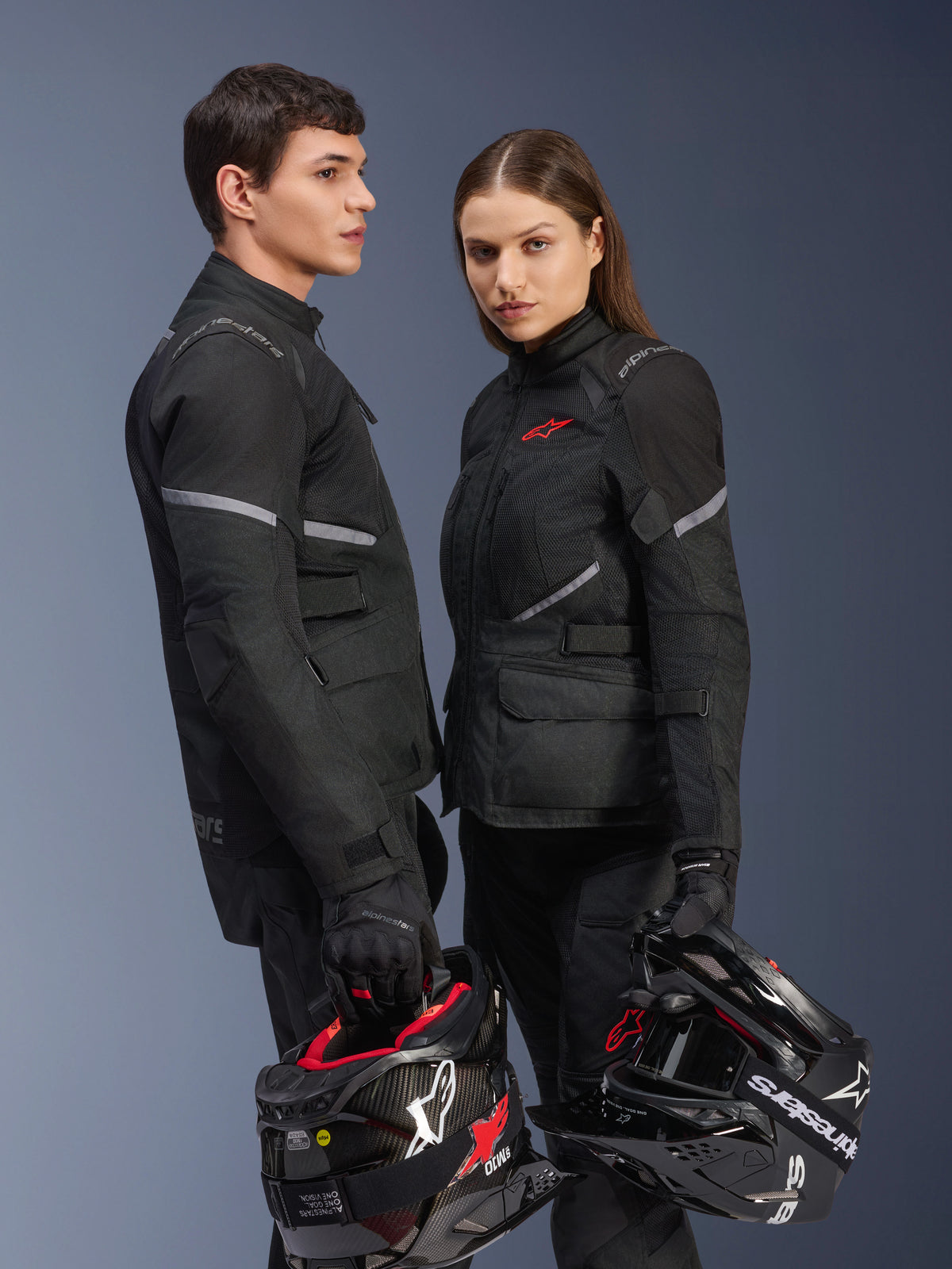Motocyklista i motocyklistka w odzieży motocyklowej Alpinestars, kurtka Stella Andes Air Drystar® w kolorze czarnym, dopasowane tekstylne spodnie i rękawice, oboje trzymają czarno-białe kaski, zdjęcie studyjne prezentujące zestaw odzieży adventure touring