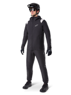 Alpinestars A-Dura Rain Jacket, Black Mountain Bike Rain Jacket, featuring white shoulder accents, worn by a rider with matching black MTB helmet, sunglasses, gloves, and pants, technical all-weather cycling outfit