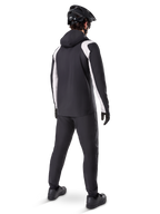 Alpinestars A-Dura Rain Jacket, back view of a male cyclist wearing the black and white waterproof MTB jacket, matching black cycling pants, black helmet, and gloves, technical gear for mountain biking in wet conditions.