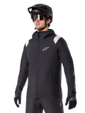 Alpinestars A-Dura Rain Jacket, Mountain Bike Rain Jacket, Black with light gray shoulder accents, worn by a rider with a black MTB helmet and sunglasses, white chest logo, waterproof construction, coordinated with black pants and gloves for off-road cycling