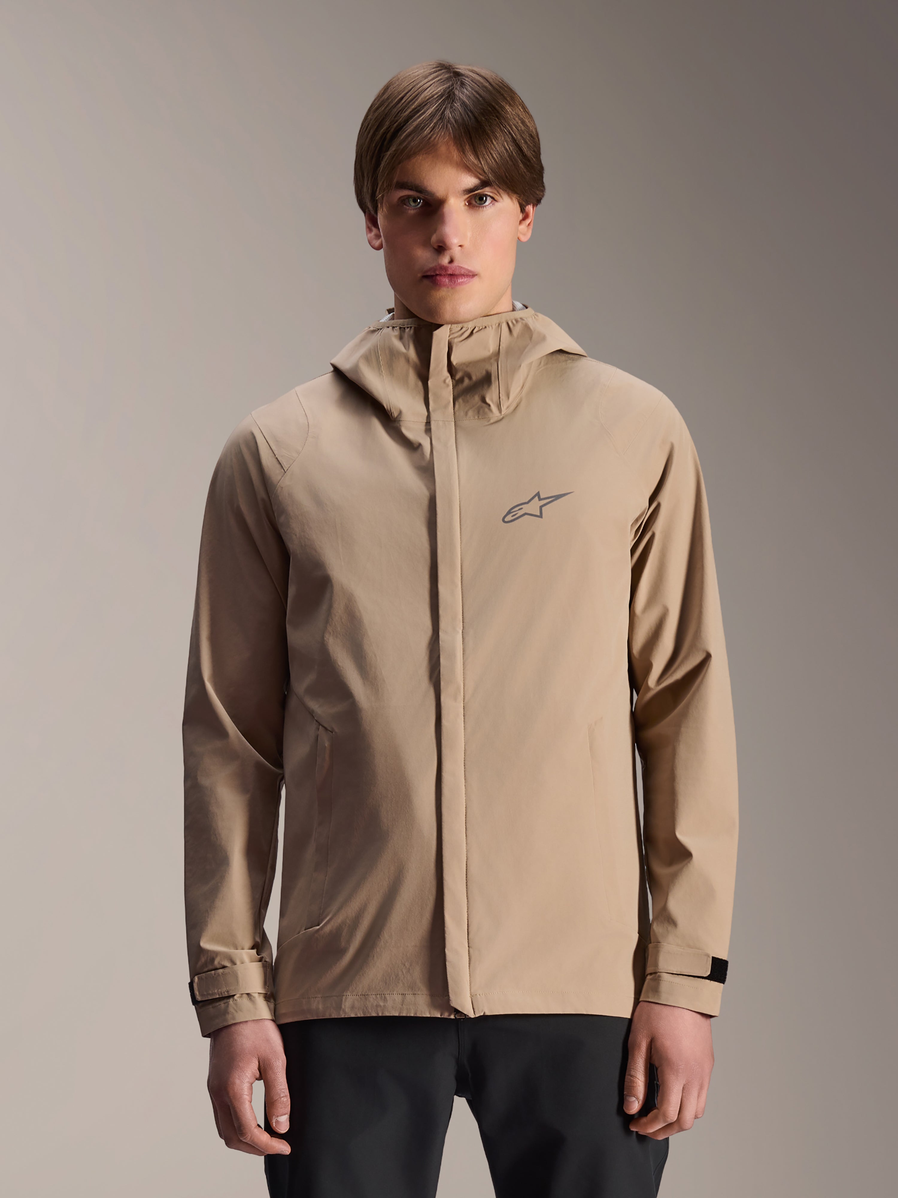 Alpinestars A-Dura Rain Jacket, jasnobrązowa kurtka przeciwdeszczowa z kapturem na rower górski, prezentowana przez modela, małe logo na piersi, regulowane paski przy mankietach i ukryty przedni zamek błyskawiczny, wodoodporna odzież rowerowa