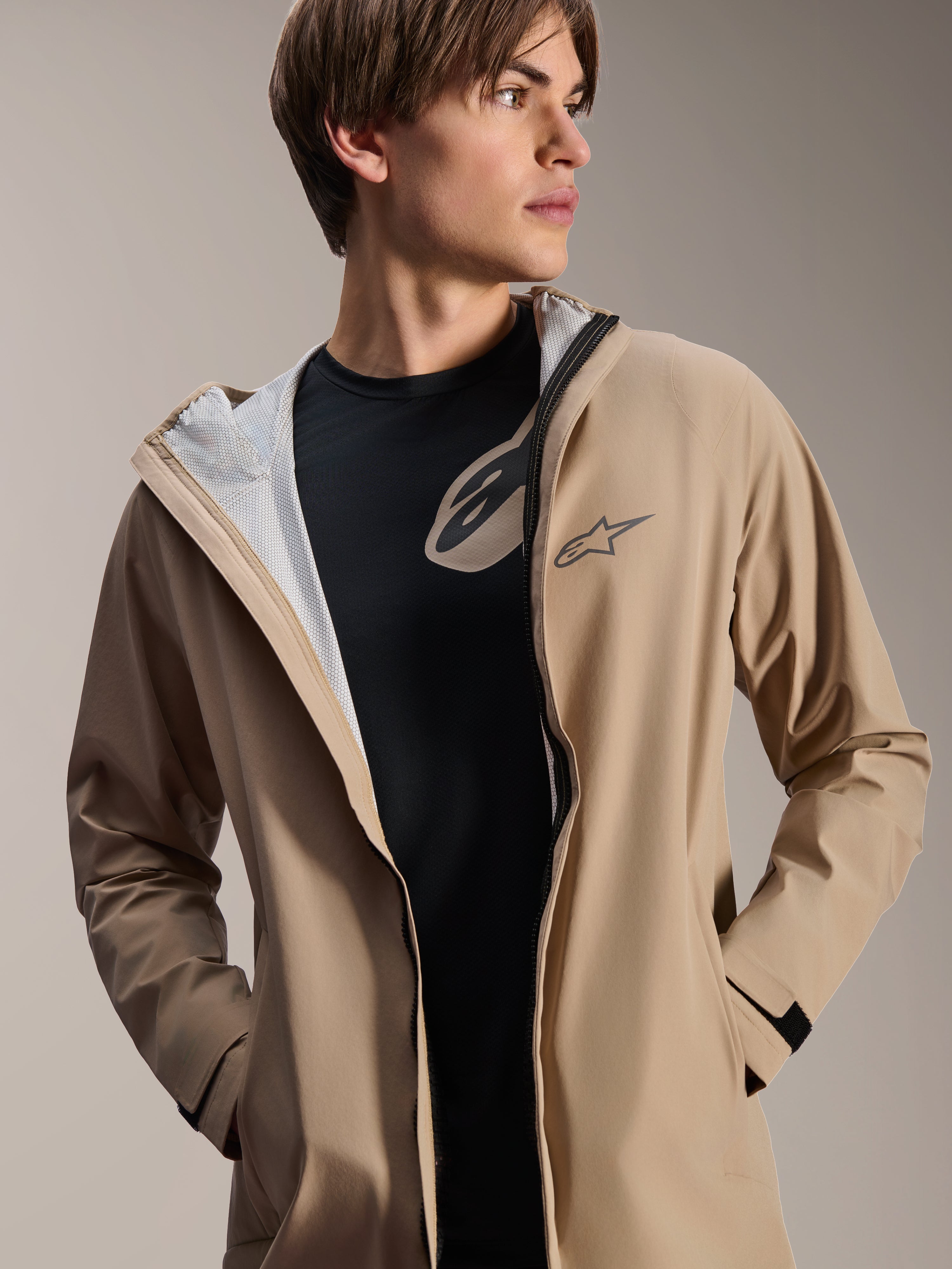 Męski model mający na sobie kurtkę przeciwdeszczową Alpinestars A-Dura w kolorze jasnobrązowym, rozpiętą na czarnym t-shircie, widoczna biała siateczkowa podszewka, wszechstronna kurtka rowerowa odporna na warunki atmosferyczne