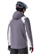 Alpinestars A-Dura Rain Jacket, kurtka przeciwdeszczowa MTB, ciemnoszara (Dark Titanium), widok z tyłu z jasnoszarymi panelami na ramionach i rękawach, czerwone logo z boku, noszona z szarym kaskiem MTB i czarno-czerwonymi rękawicami, techniczna odzież trailowa
