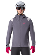 Kurtka przeciwdeszczowa Alpinestars A-Dura, rowerowa kurtka przeciwdeszczowa MTB, kolor ciemny tytanowy szary, fason z kapturem z różowym logo Alpinestars i jasnoszarymi panelami na ramionach, noszona z białym kaskiem MTB i jaskraworóżowymi rękawiczkami, wodoodporna odzież do jazdy w terenie