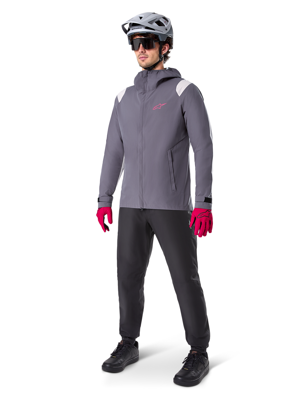 Model w kurtce przeciwdeszczowej Alpinestars A-Dura w kolorze Dark Titanium, szara kurtka z kapturem z różowym logo na piersi i jasnoszarymi panelami na ramionach, zestawiona z szarym kaskiem MTB, ciemnymi okularami, jaskraworóżowymi rękawicami i czarnymi spodniami do jazdy terenowej.