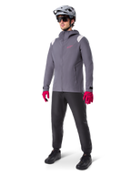 Model w kurtce przeciwdeszczowej Alpinestars A-Dura w kolorze Dark Titanium, szara kurtka z kapturem z różowym logo na piersi i jasnoszarymi panelami na ramionach, zestawiona z szarym kaskiem MTB, ciemnymi okularami, jaskraworóżowymi rękawicami i czarnymi spodniami do jazdy terenowej.