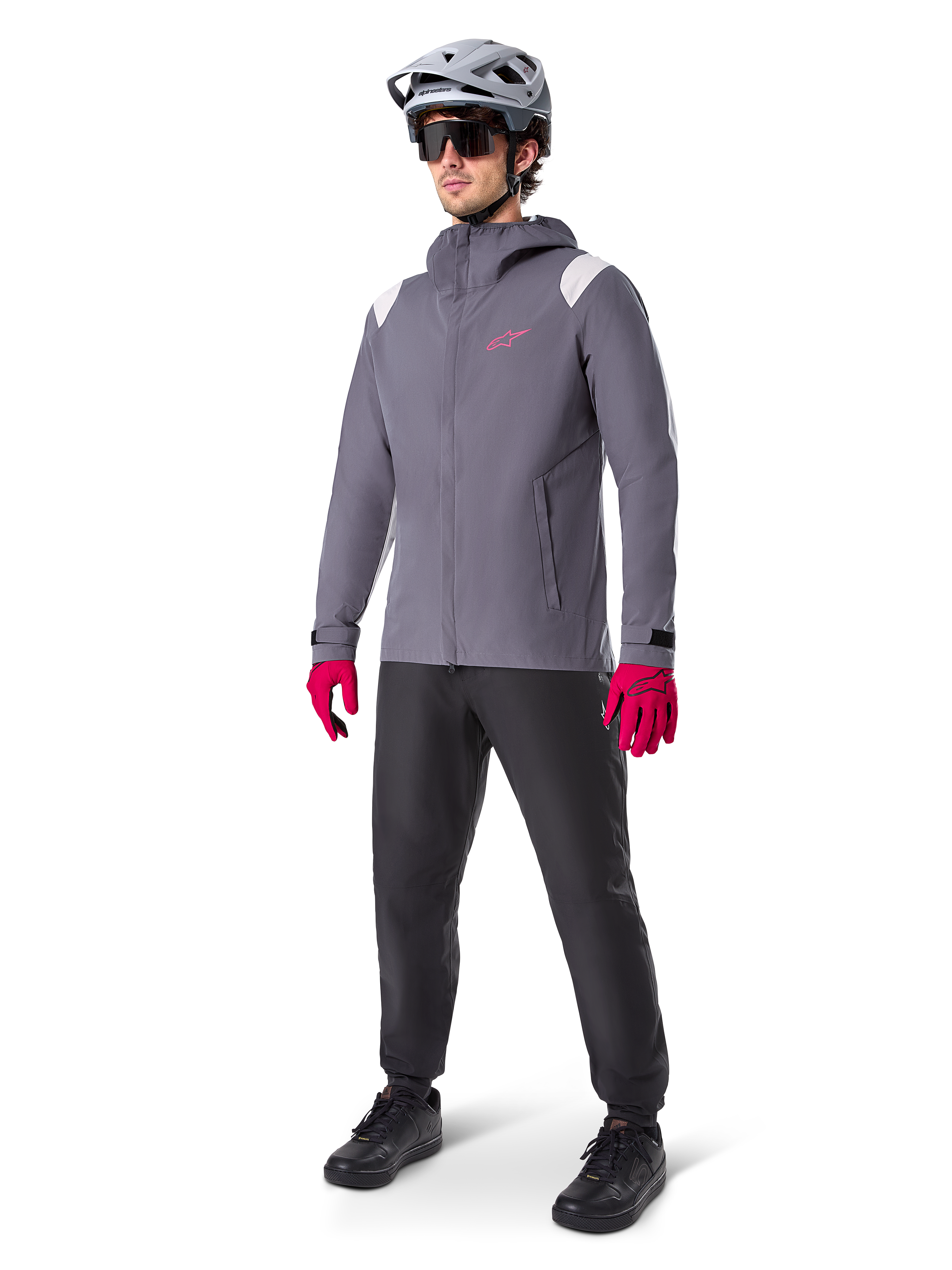 Model w kurtce przeciwdeszczowej Alpinestars A-Dura w kolorze Dark Titanium, szara kurtka z kapturem z różowym logo na piersi i jasnoszarymi panelami na ramionach, zestawiona z szarym kaskiem MTB, ciemnymi okularami, jaskraworóżowymi rękawicami i czarnymi spodniami do jazdy terenowej.