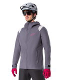 Alpinestars A-Dura Rain Jacket, rowerowa kurtka przeciwdeszczowa MTB, ciemnoszara z różowym logo i białymi akcentami na ramionach, model z kapturem i przednimi kieszeniami, noszona z różowymi rękawiczkami i jasnoszarym kaskiem rowerowym