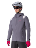 Alpinestars A-Dura Rain Jacket, rowerowa kurtka przeciwdeszczowa MTB, ciemnoszara z różowym logo i białymi akcentami na ramionach, model z kapturem i przednimi kieszeniami, noszona z różowymi rękawiczkami i jasnoszarym kaskiem rowerowym
