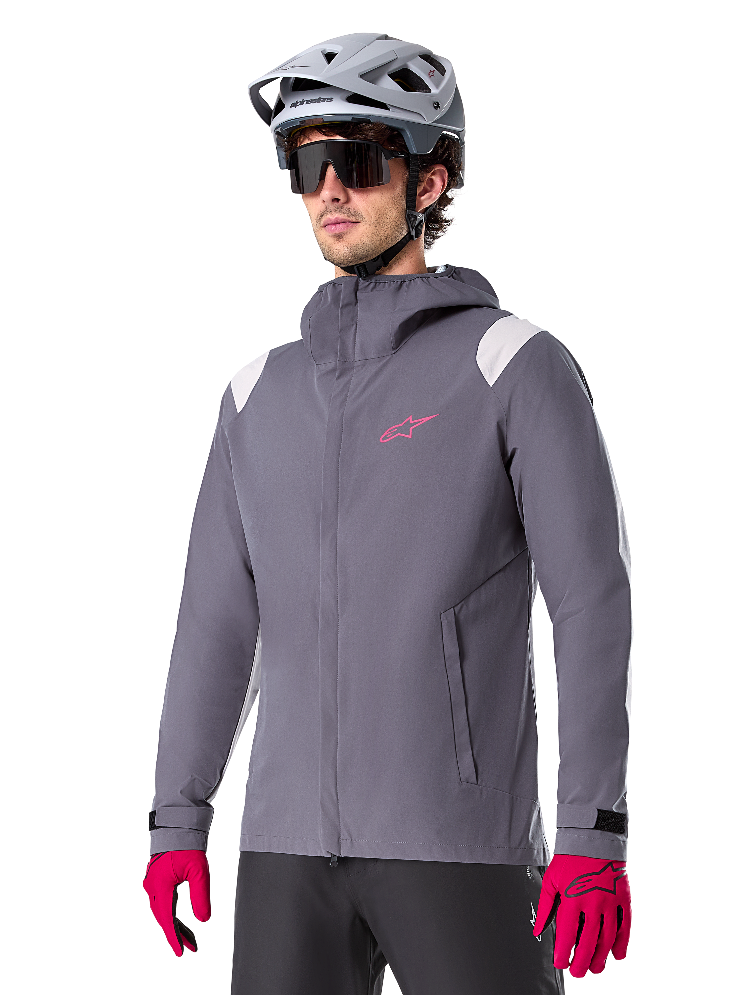 Alpinestars A-Dura Rain Jacket, rowerowa kurtka przeciwdeszczowa MTB, ciemnoszara z różowym logo i białymi akcentami na ramionach, model z kapturem i przednimi kieszeniami, noszona z różowymi rękawiczkami i jasnoszarym kaskiem rowerowym