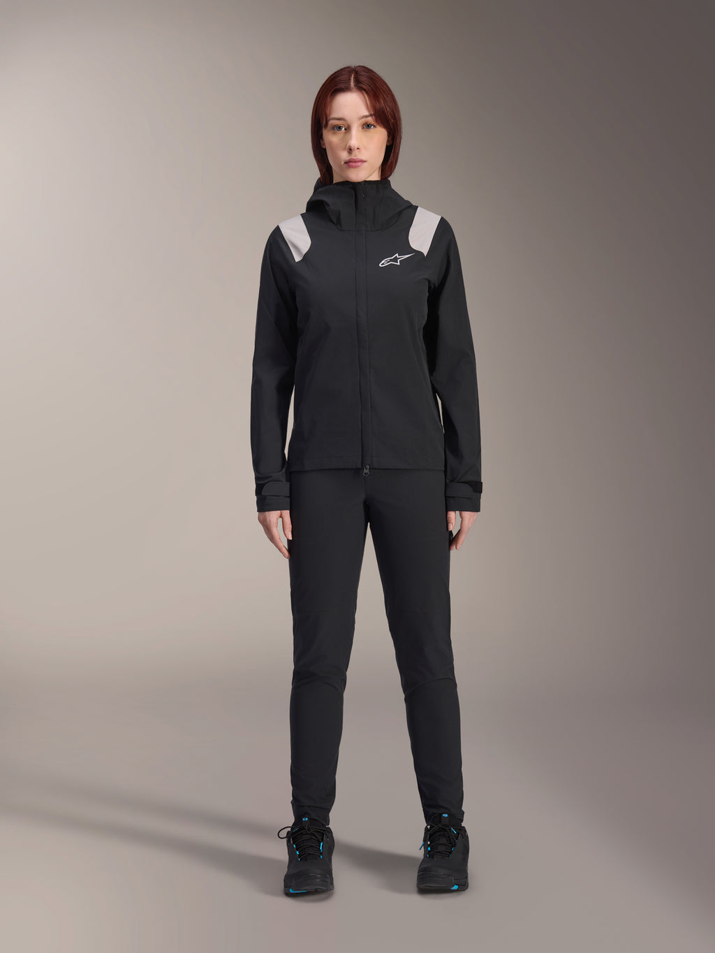 Alpinestars Stella A-Dura Rain Jacket, damska kurtka przeciwdeszczowa MTB, czarna z jasnoszarymi akcentami na ramionach i białym logo, noszona z pasującymi czarnymi spodniami i butami, wyposażona w kaptur i regulowane mankiety, zaprojektowana do jazdy na rowerze górskim