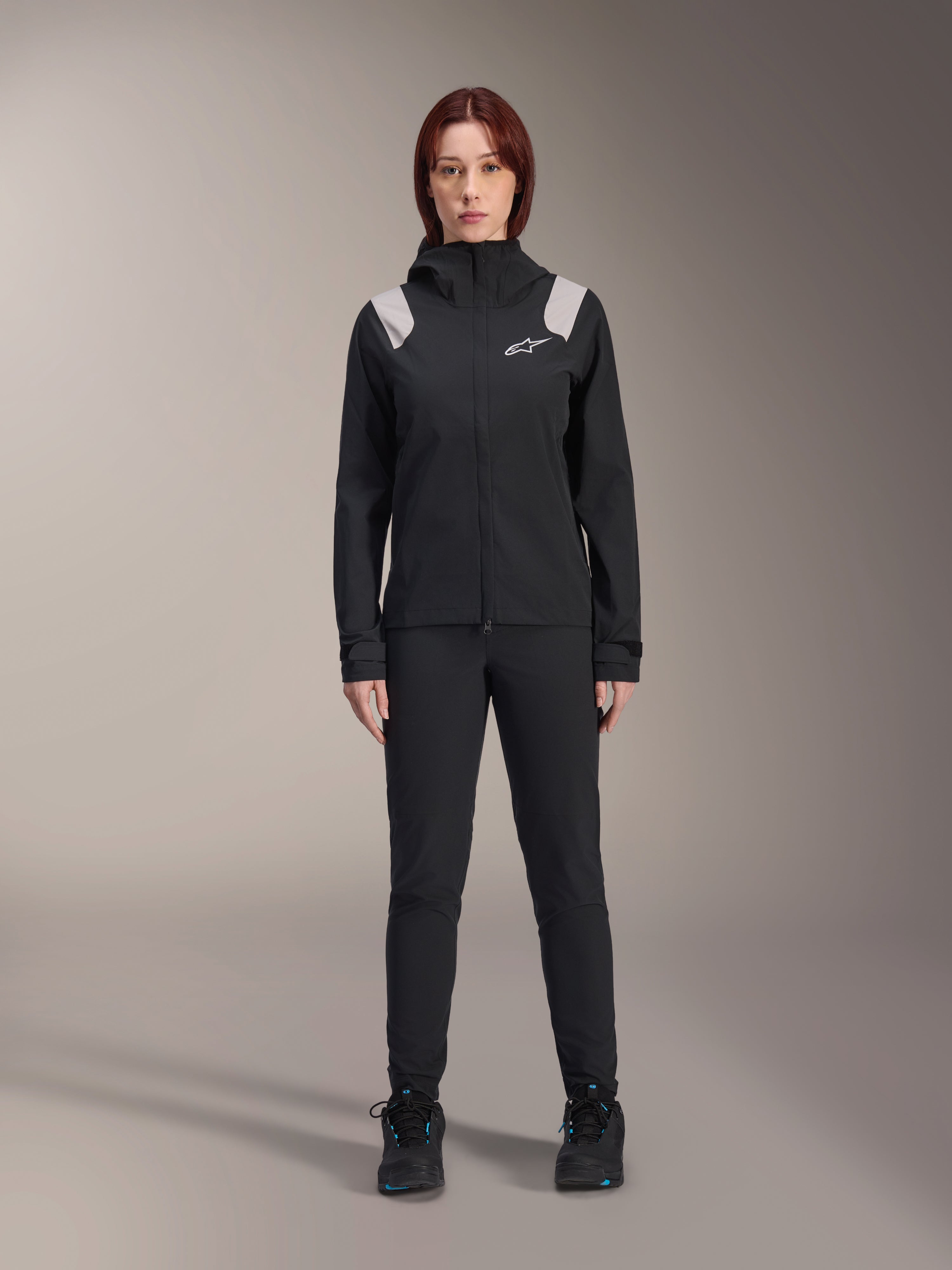 Alpinestars Stella A-Dura Rain Jacket, damska kurtka przeciwdeszczowa MTB, czarna z jasnoszarymi akcentami na ramionach i białym logo, noszona z pasującymi czarnymi spodniami i butami, wyposażona w kaptur i regulowane mankiety, zaprojektowana do jazdy na rowerze górskim