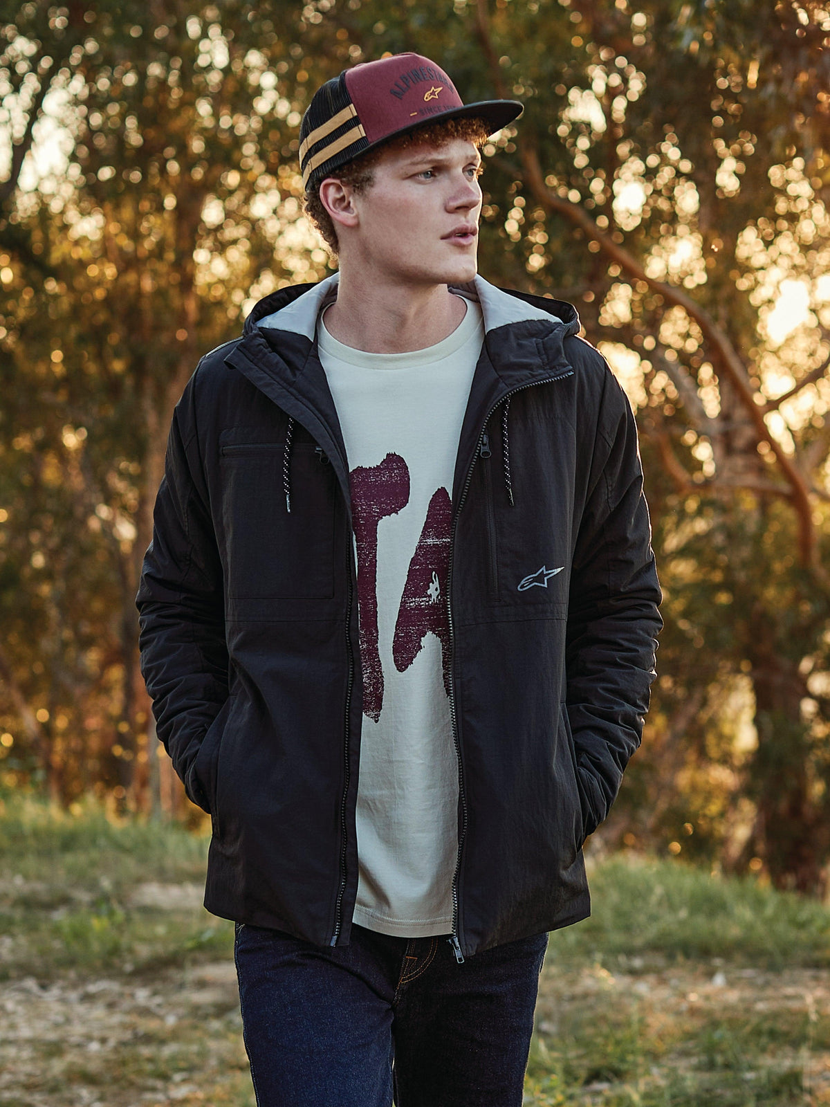 Czapka Alpinestars Backline Trucker Hat w kolorach ceglastym, czarnym i musztardowym, noszona przez mężczyznę w zestawieniu z czarną kurtką z kapturem i koszulką z grafiką, casualowa odzież uliczna inspirowana motorsportem w plenerze o złotej godzinie.