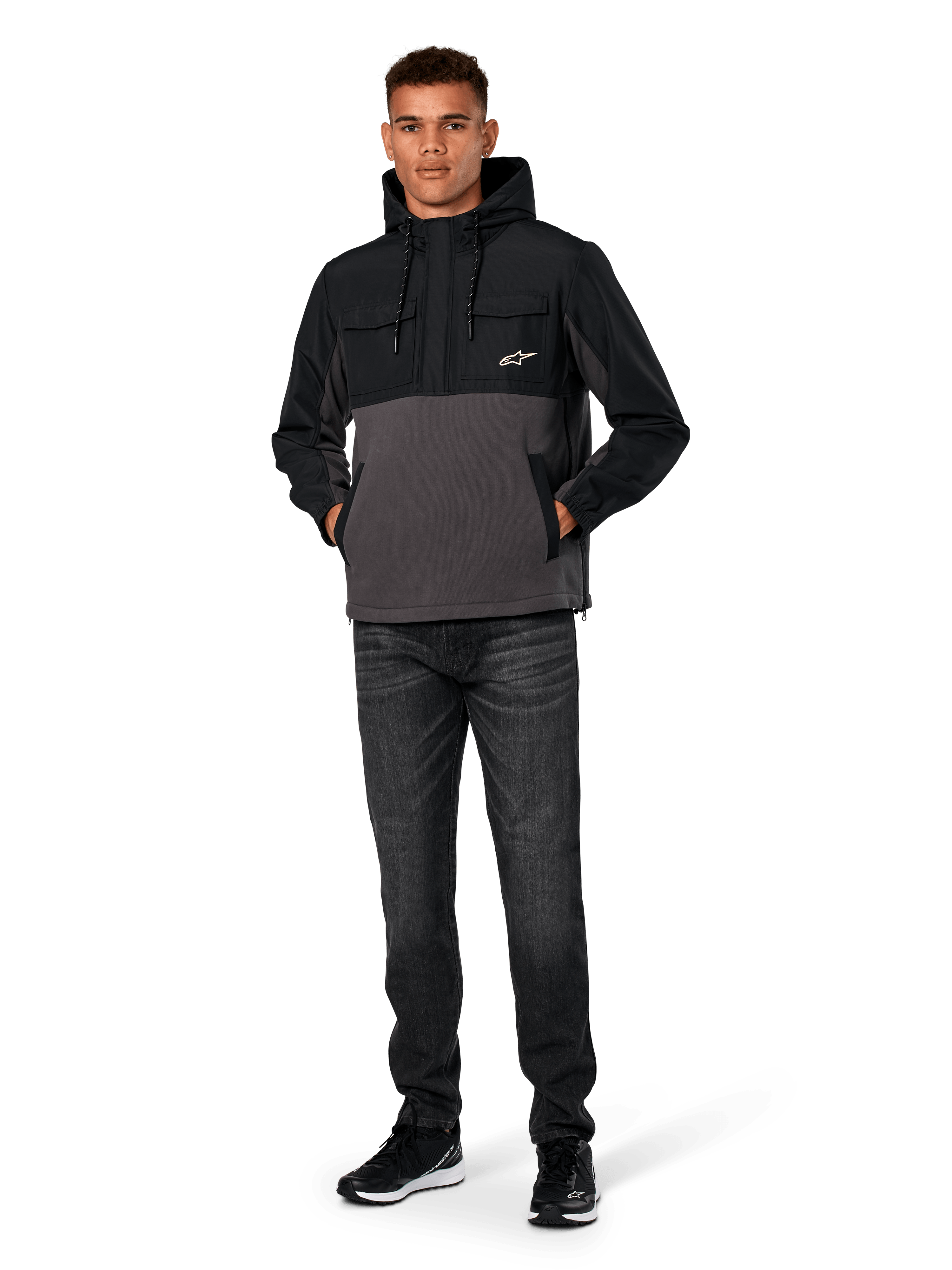 Kurtka Alpinestars Juncture Hybrid, czarno-szara (Gunmetal), anorak z kapturem z mieszanych materiałów z polarem płóciennym i nakładkami ripstop, białe logo A-star na piersi, kaptur ze sznurkiem, model w ciemnych dżinsach i czarnych sneakersach, codzienna odzież uliczna inspirowana motorsportem