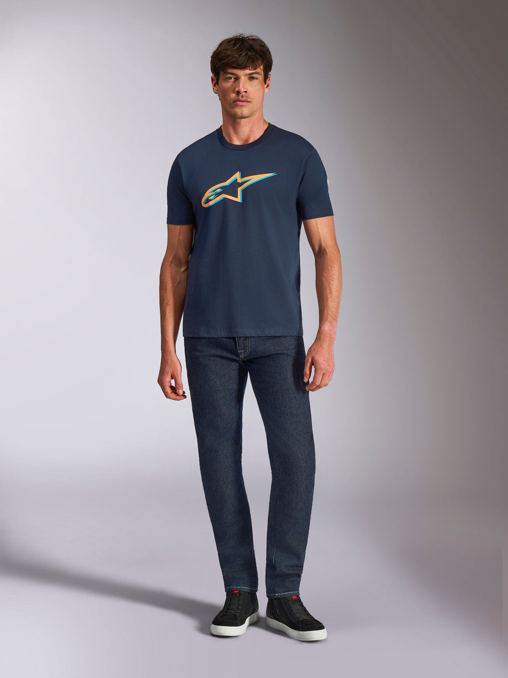 Alpinestars Ageless Shadow CSF Tee, t-shirt z krótkim rękawem, granatowy, z pomarańczowym i jasnoniebieskim logo Ageless Shadow, prezentowany na modelu z ciemnymi dżinsami i czarnymi sneakersami, codzienny styl streetwearowy inspirowany motorsportem