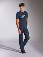 Alpinestars Ageless Shadow CSF Tee, koszulka z krótkim rękawem, granatowa z pomarańczowym i turkusowym logo Shadow, prezentowana przez modela w ciemnych jeansach i czarnych sneakersach, codzienna odzież typu motorsport