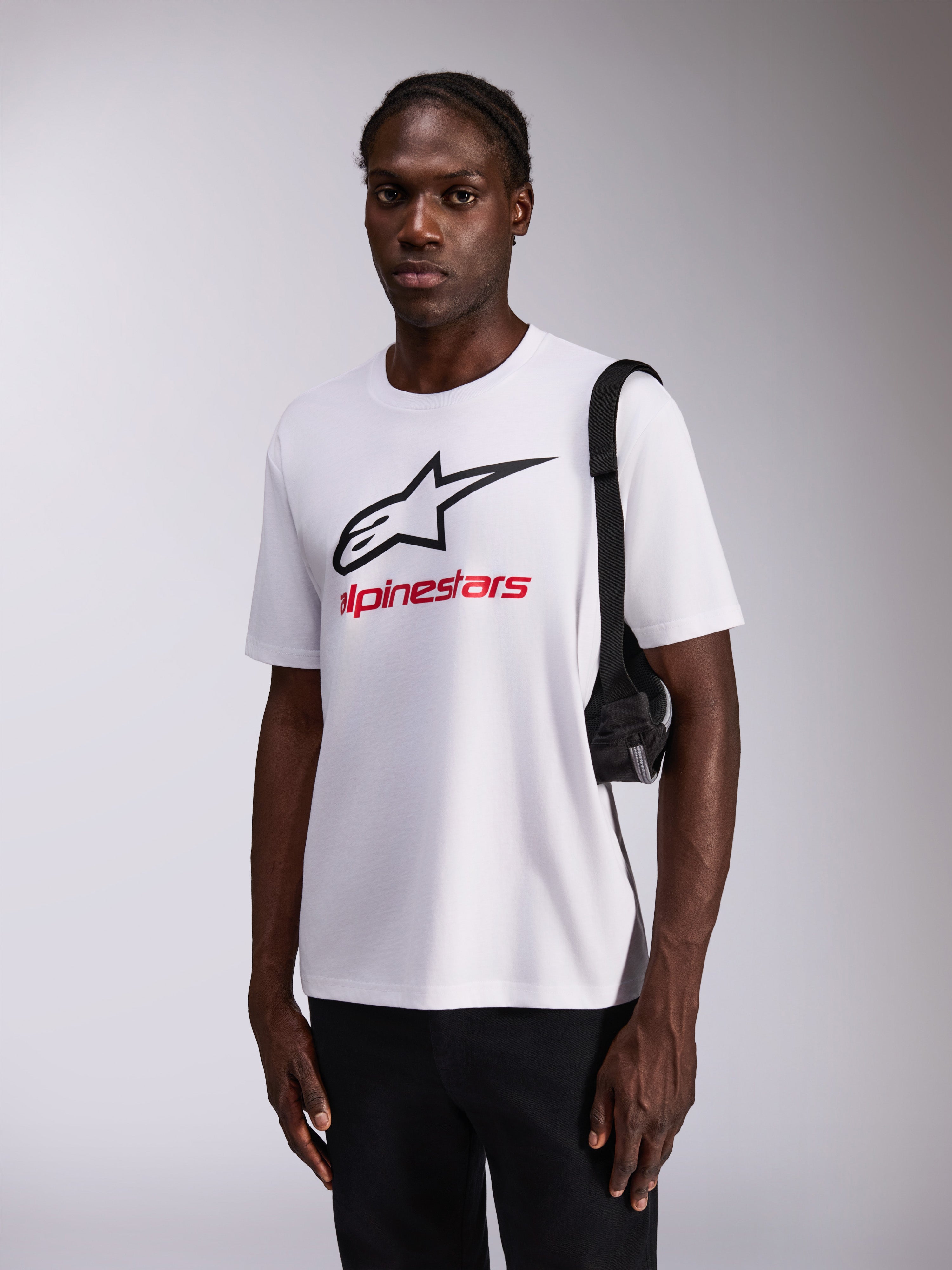 Koszulka Alpinestars Always 2.0 CSF, T-shirt z krótkim rękawem, biała z dużym czarnym logo gwiazdy i czerwonym brandingiem, model w czarnych spodniach z torbą na ramię, casualowy streetwear inspirowany sportami motorowymi