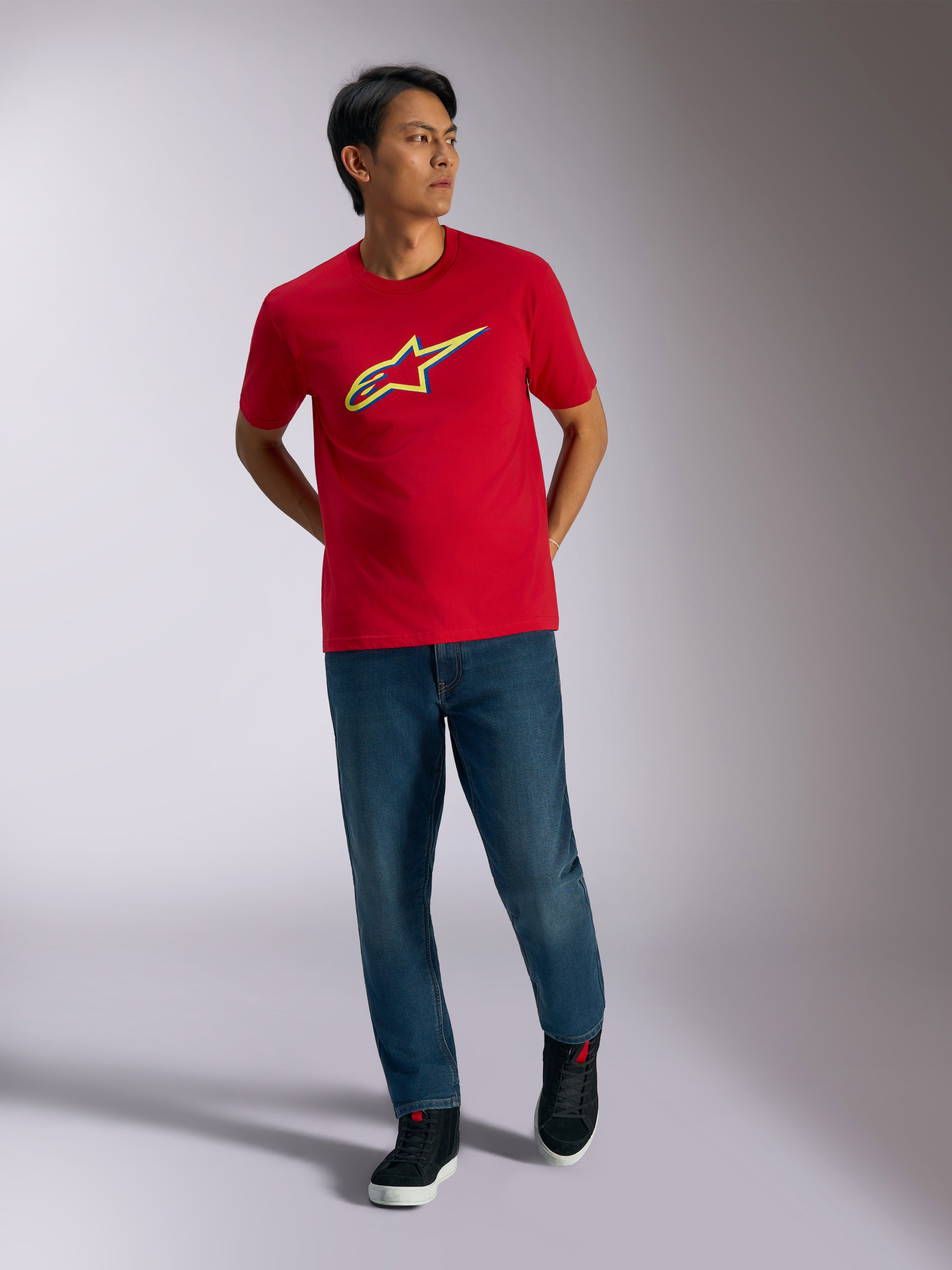 Alpinestars Ageless Shadow CSF Tee, czerwona koszulka z krótkim rękawem, żółto-niebieska grafika z logo shadow, noszona z niebieskimi jeansami i czarnymi sneakersami, codzienna odzież lifestylowa motorsport
