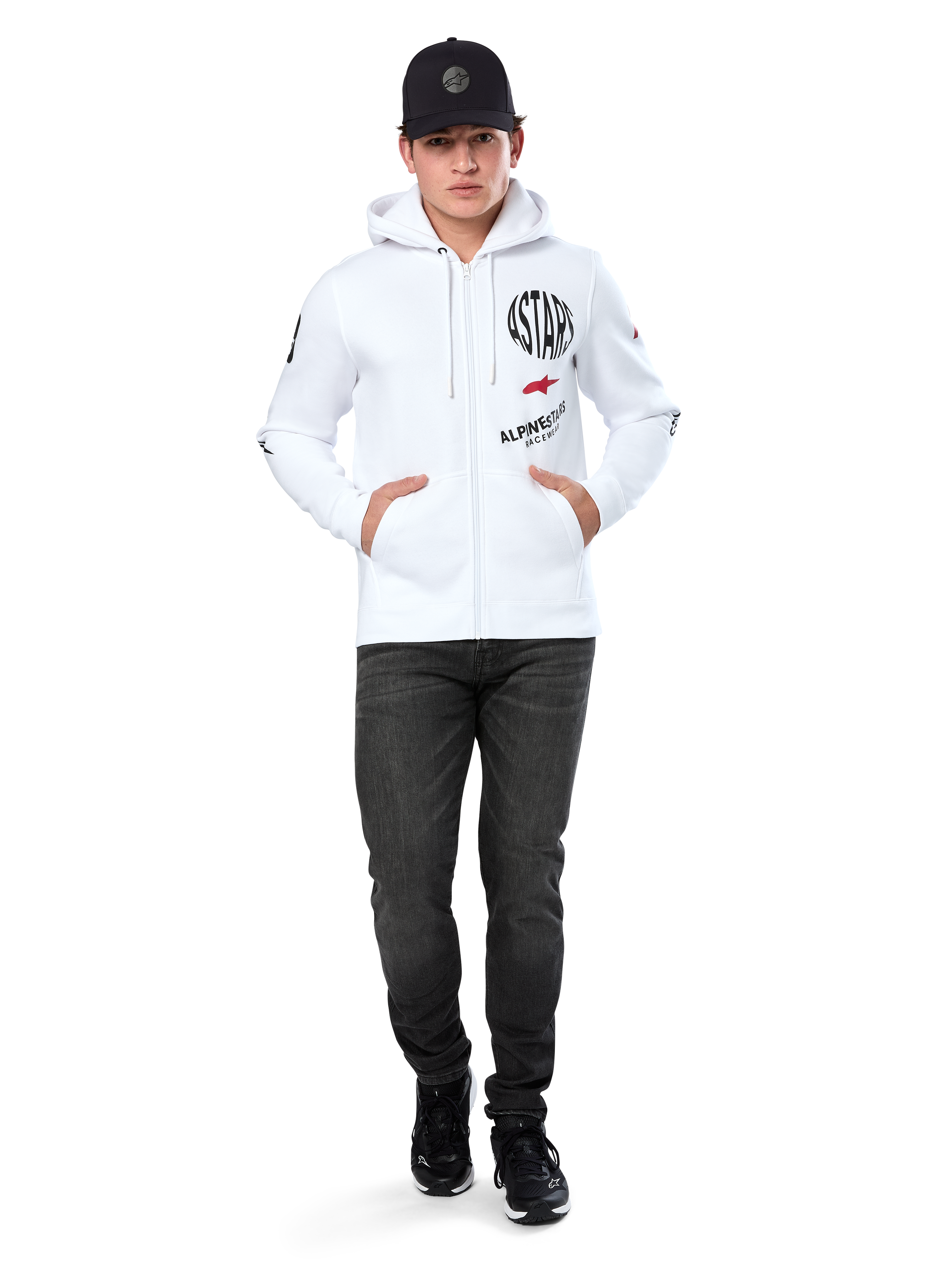 Bluza z kapturem Alpinestars Variegate, biała rozpinana bluza polarowa z kapturem, wieloelementowa grafika z napisem „ASTARS” i czerwonym logo gwiazdy na piersi, detale graficzne na rękawach, model w czarnej czapce z daszkiem i czarnych jeansach, codzienna odzież streetwear w stylu motorsport