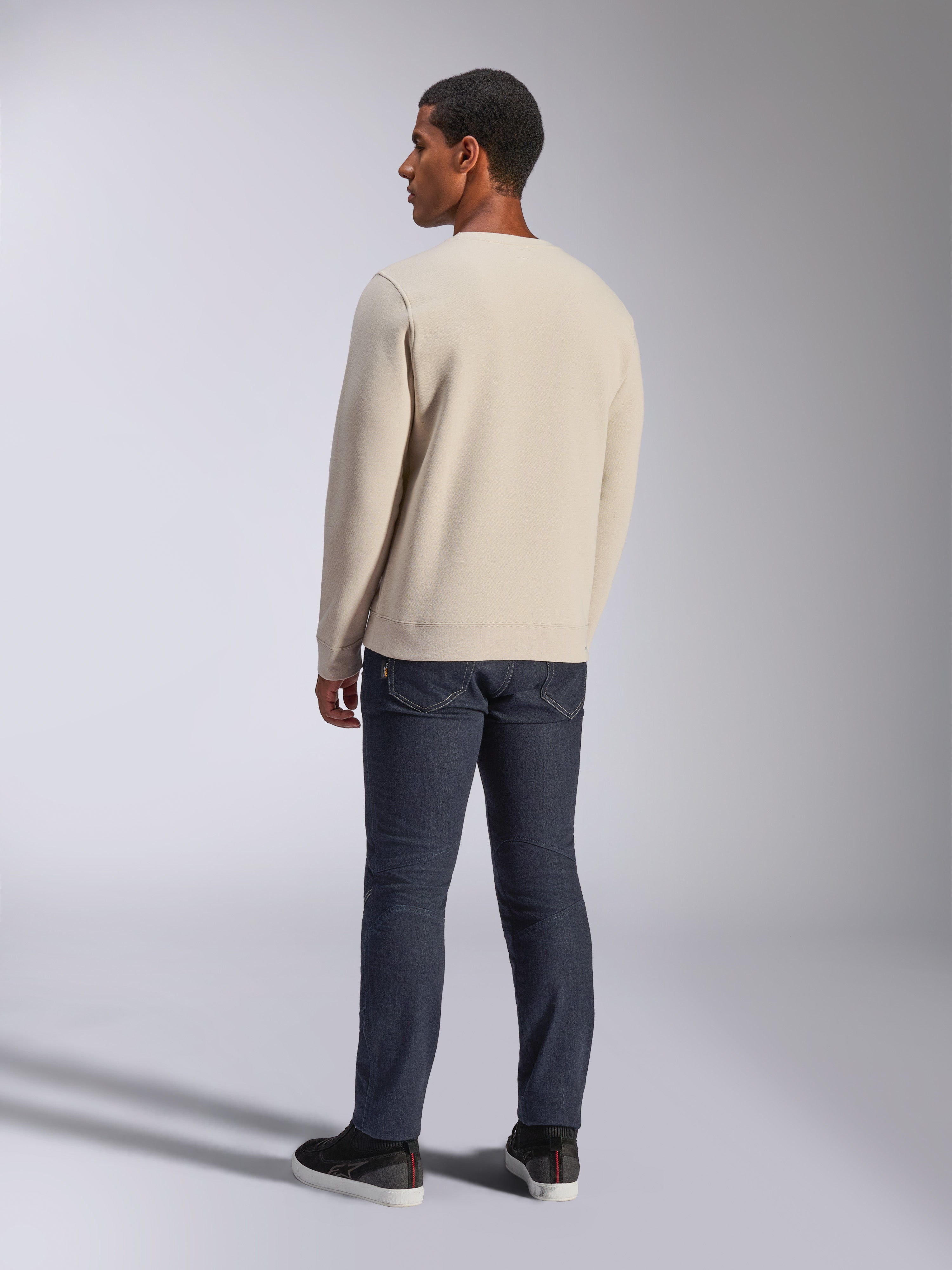 Alpinestars Positive Crew, bluza typu crewneck w kolorze złamanej bieli, widok z tyłu na modelu, w zestawieniu z ciemnymi jeansami i czarnymi sneakersami, casualowa odzież uliczna w stylu motorsport, ze ściągaczami przy mankietach i na dole