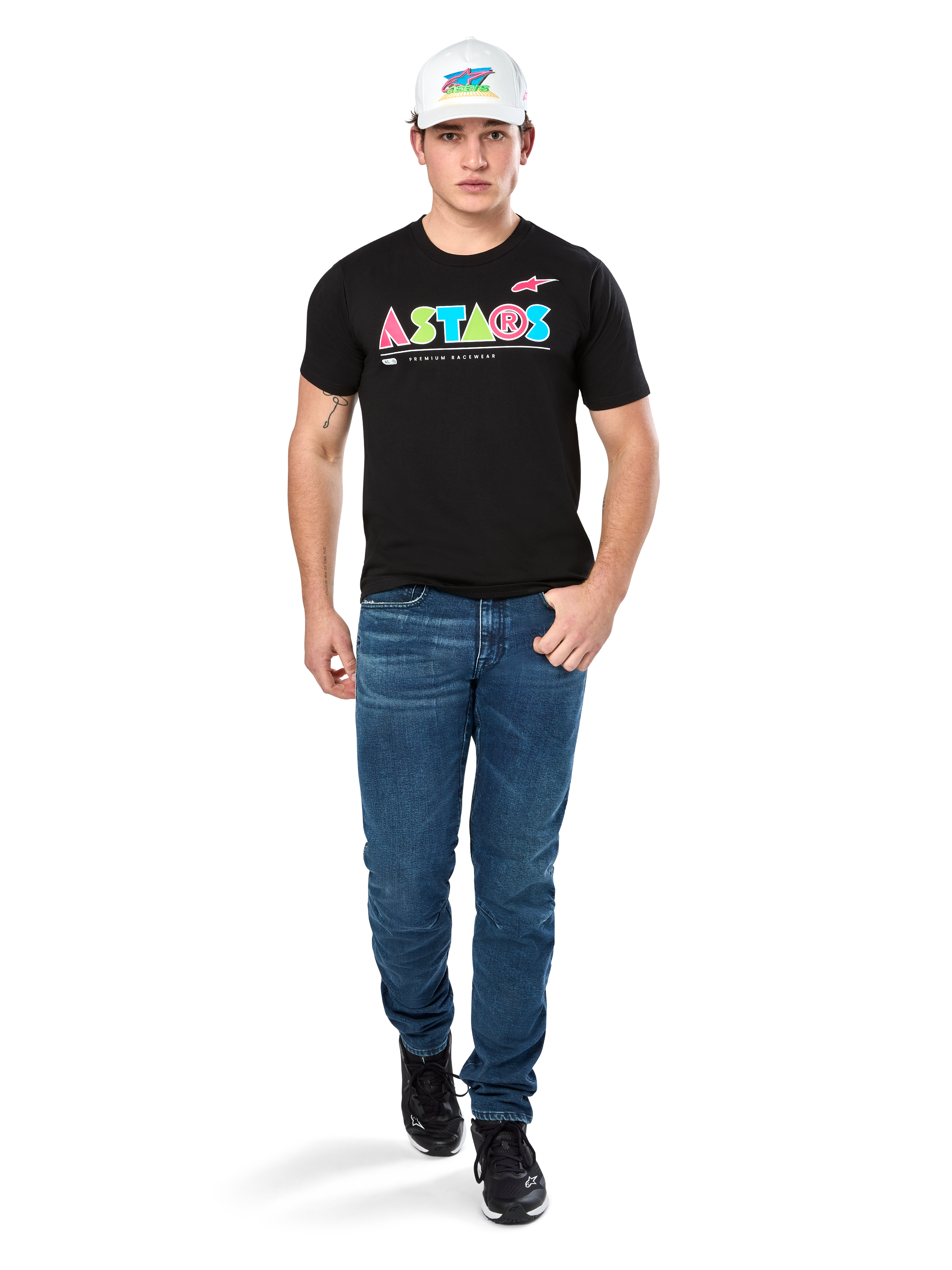 Męski model ubrany w koszulkę Alpinestars Cereal CSF Tee, czarny T-shirt z krótkim rękawem z kolorowym geometrycznym logo ASTARS, zestawiony z niebieskimi jeansami i białą czapką, casualowy styl streetwear inspirowany motorsportem