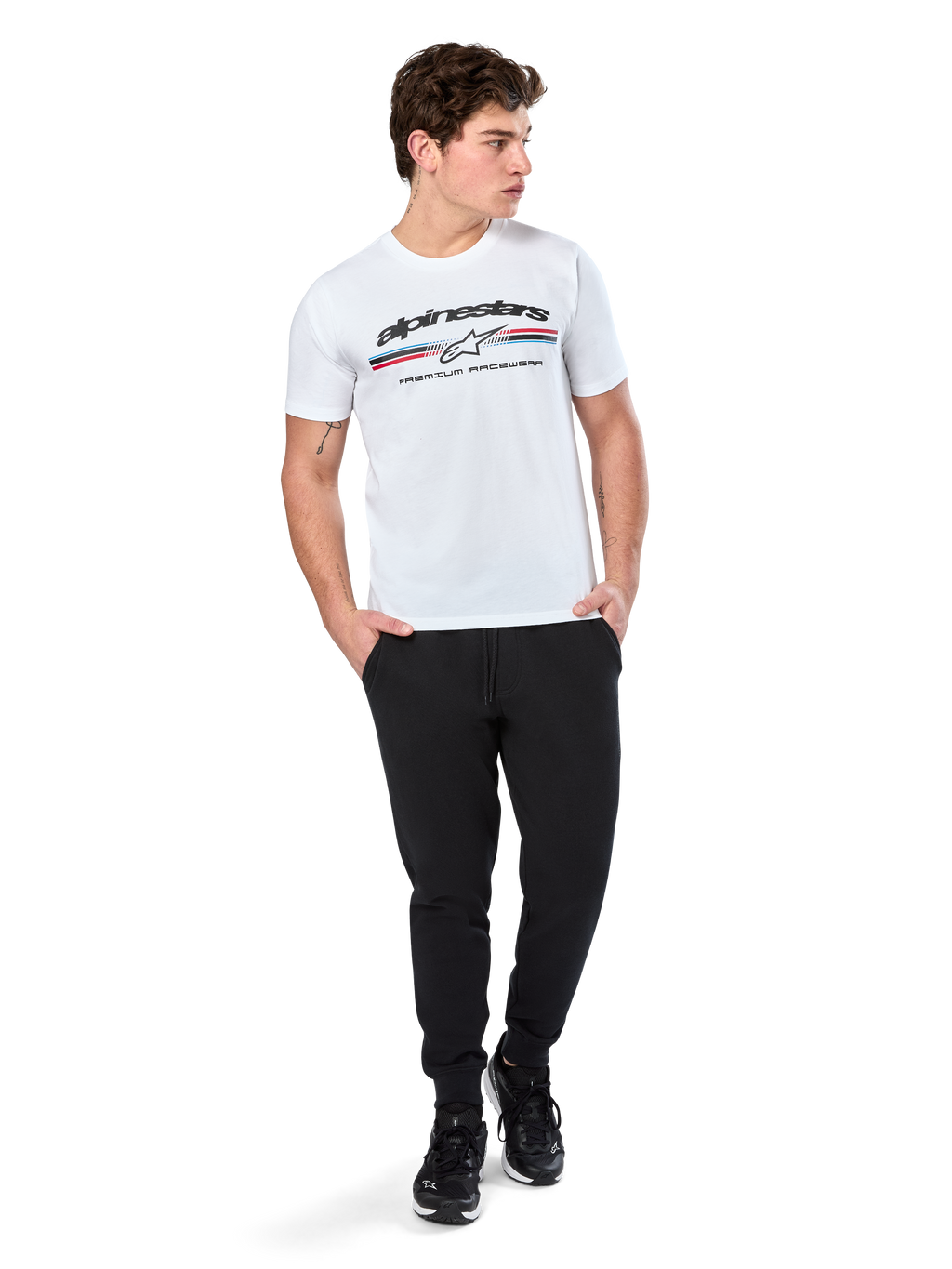 Alpinestars Prevail CSF Tee, koszulka z krótkim rękawem, biała, z logo z trzema kolorowymi paskami w stylu paddock i brandingiem Alpinestars z przodu, prezentowana na modelu w czarnych joggerach i czarnych sneakersach, codzienna moda uliczna w stylu motorsport