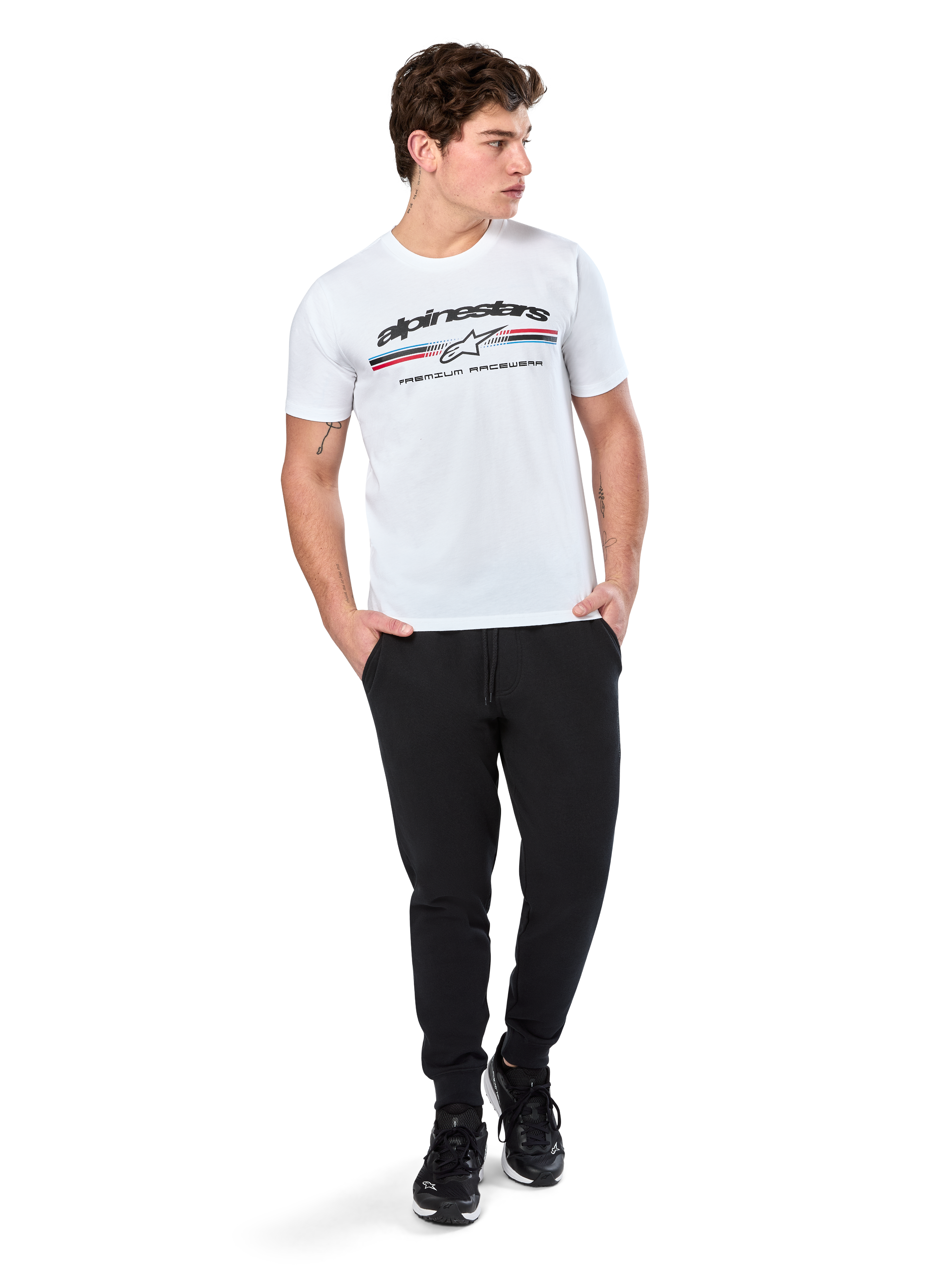 Alpinestars Prevail CSF Tee, koszulka z krótkim rękawem, biała, z logo z trzema kolorowymi paskami w stylu paddock i brandingiem Alpinestars z przodu, prezentowana na modelu w czarnych joggerach i czarnych sneakersach, codzienna moda uliczna w stylu motorsport
