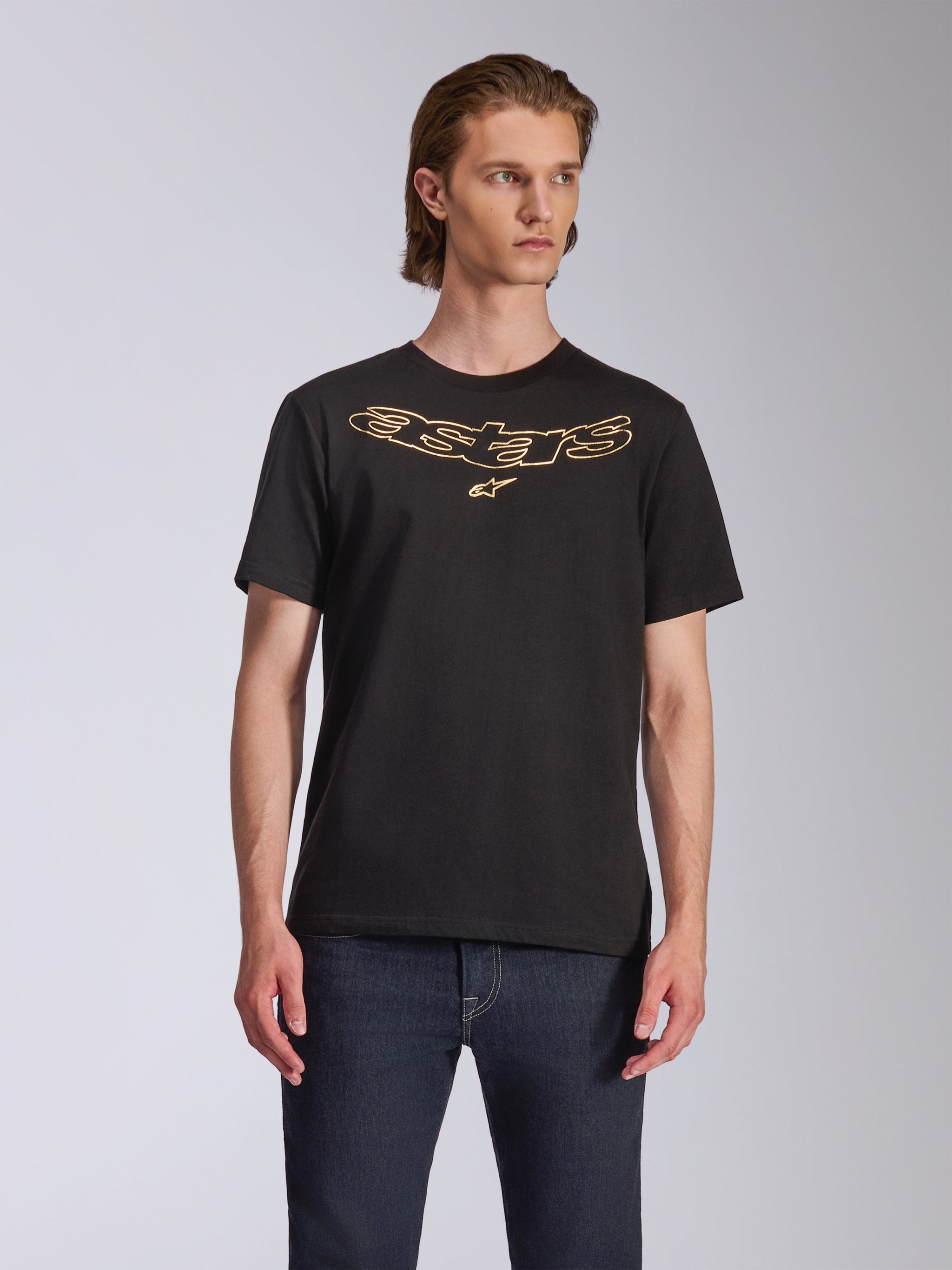 Alpinestars Luxo CSF Tee, koszulka z krótkim rękawem, czarna, złote konturowe logo Astars na piersi, noszona z ciemnoniebieskimi dżinsami, casualowy styl motorsportowy