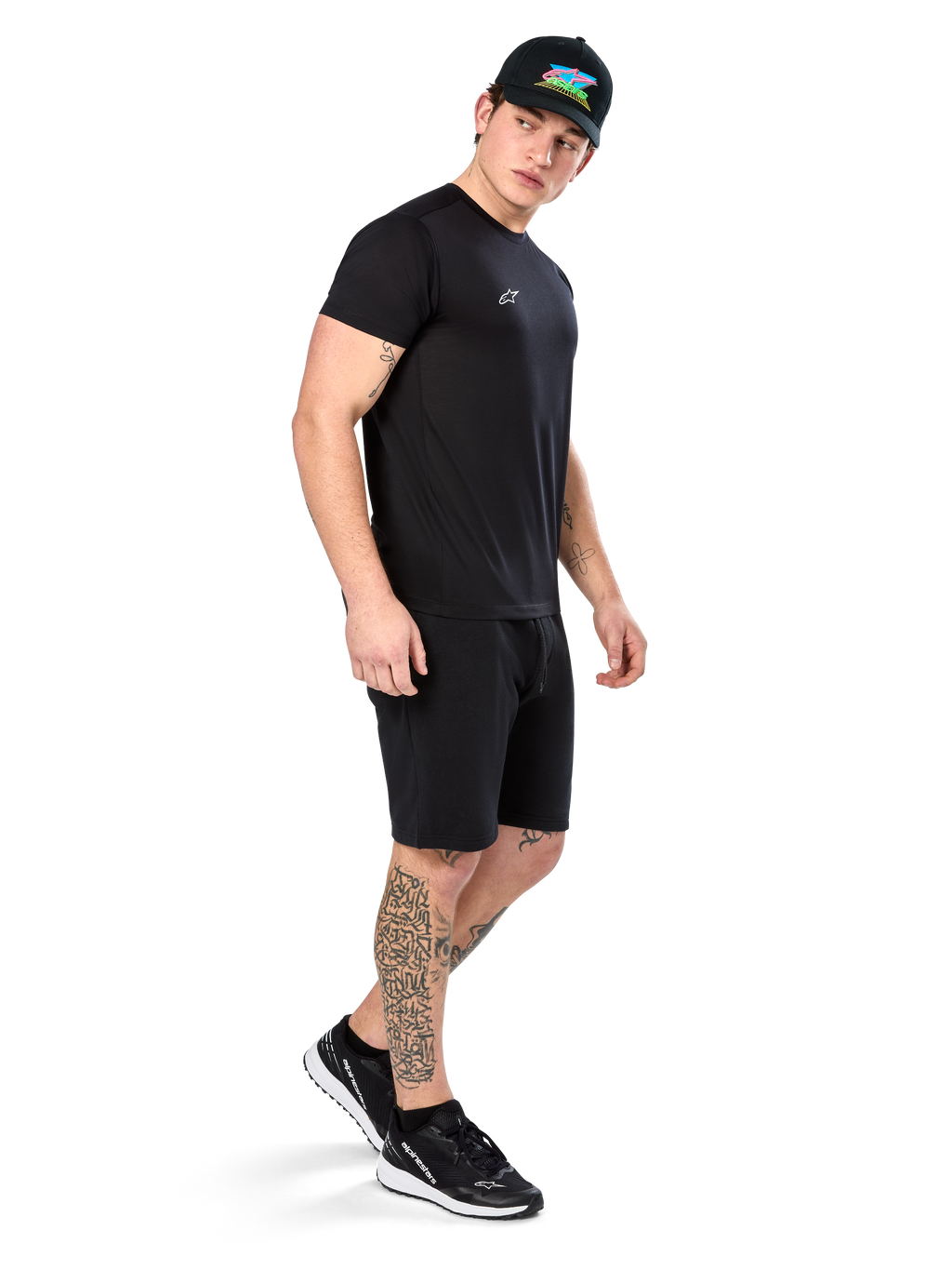 Mężczyzna z tatuażami w koszulce Alpinestars Point Performance Tee, czarny t-shirt techniczny z krótkim rękawem, małe białe logo, w zestawie z czarnymi szortami, czarną czapką z kolorową grafiką i czarno-białymi sneakersami, sportowy styl lifestyle